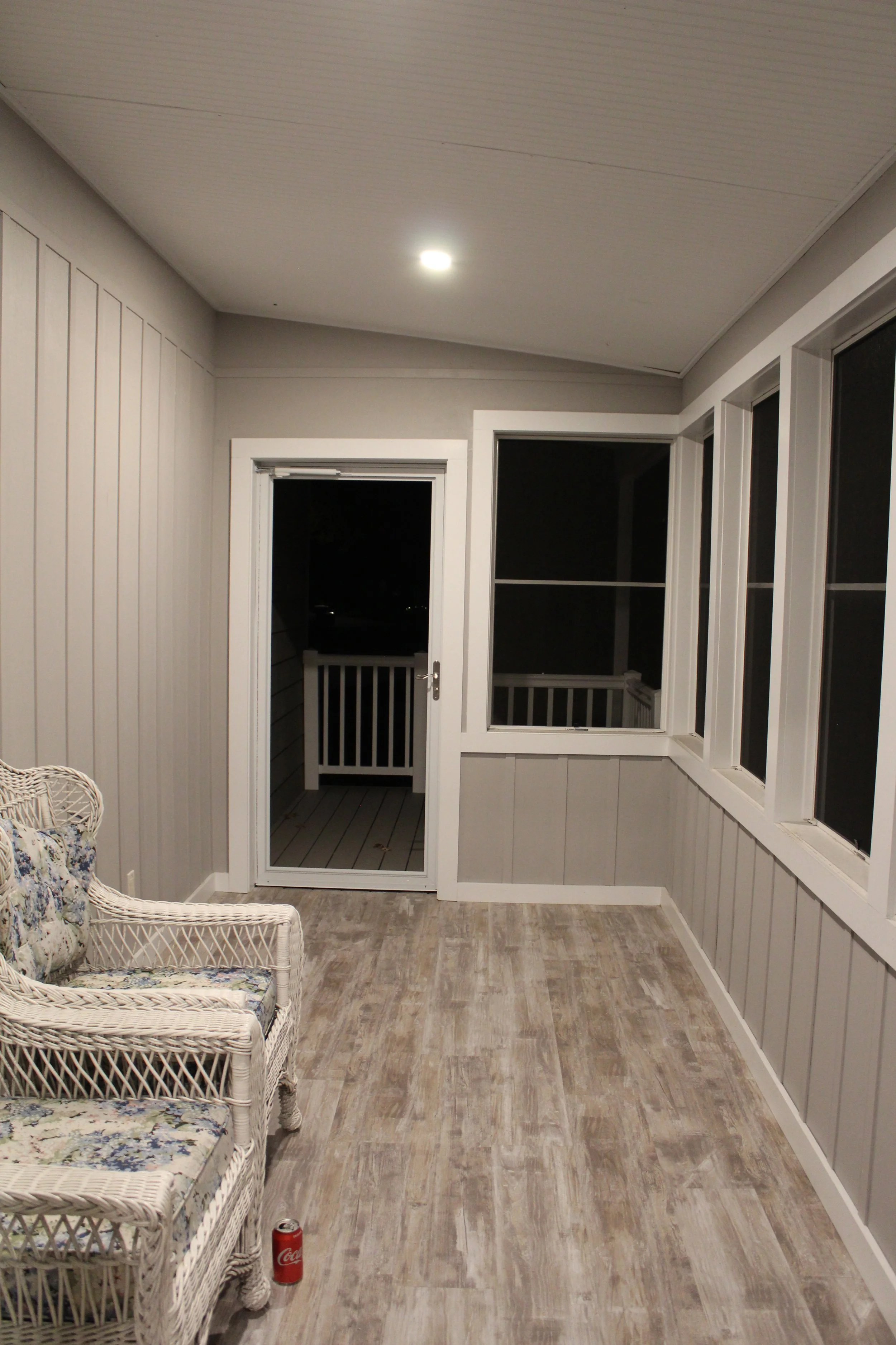 A screened-in porch at night with a wicker couch, a ceiling light, a glass door leading outside, and several windows.