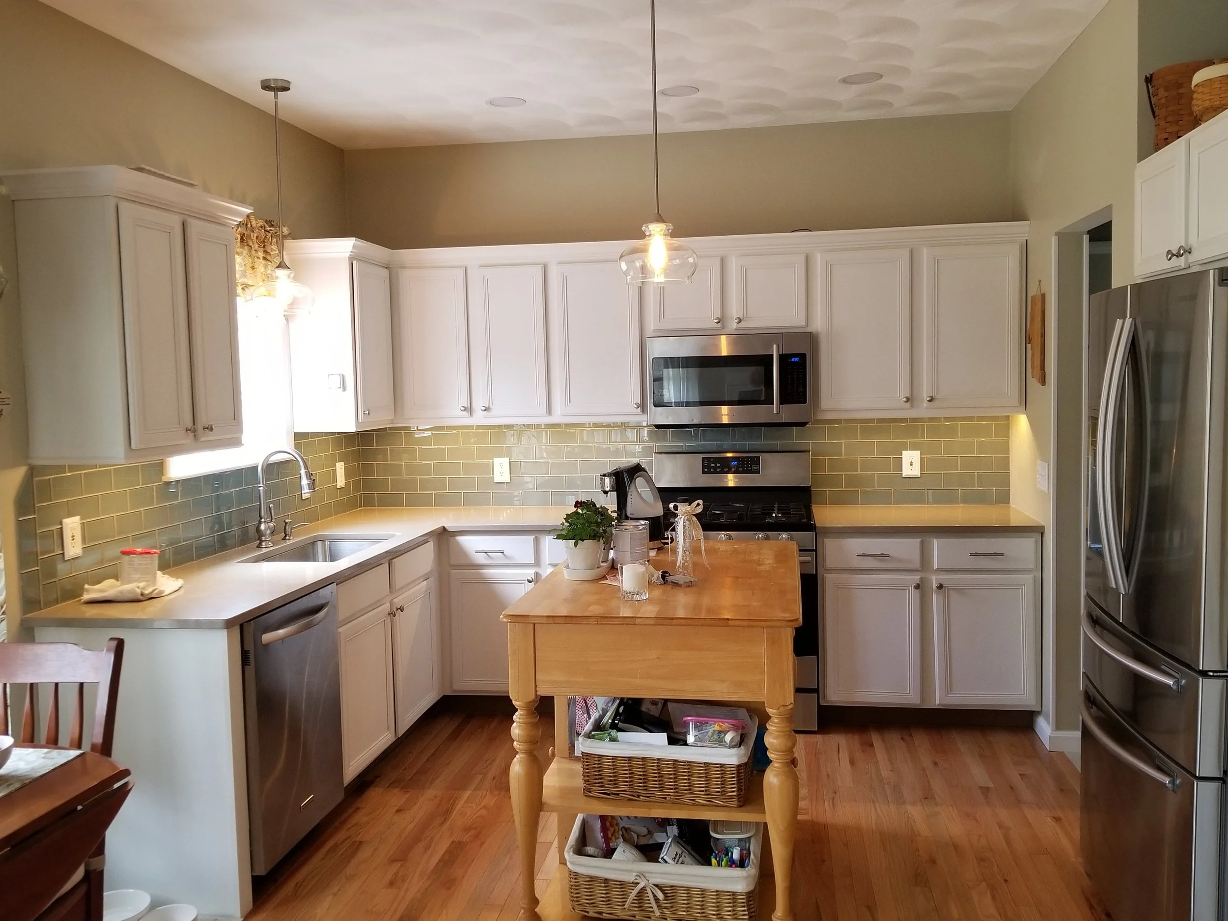Kitchen with white cabinets, stainless steel appliances, a small wooden island, green tile backsplash, hardwood floors, and a window with a floral valance.