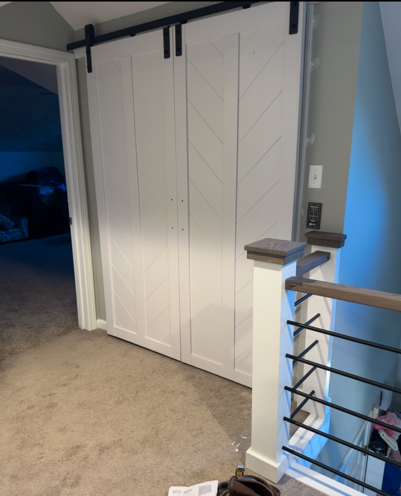 White sliding barn door on black track inside a house, next to staircase with wooden handrail and black metal bars.