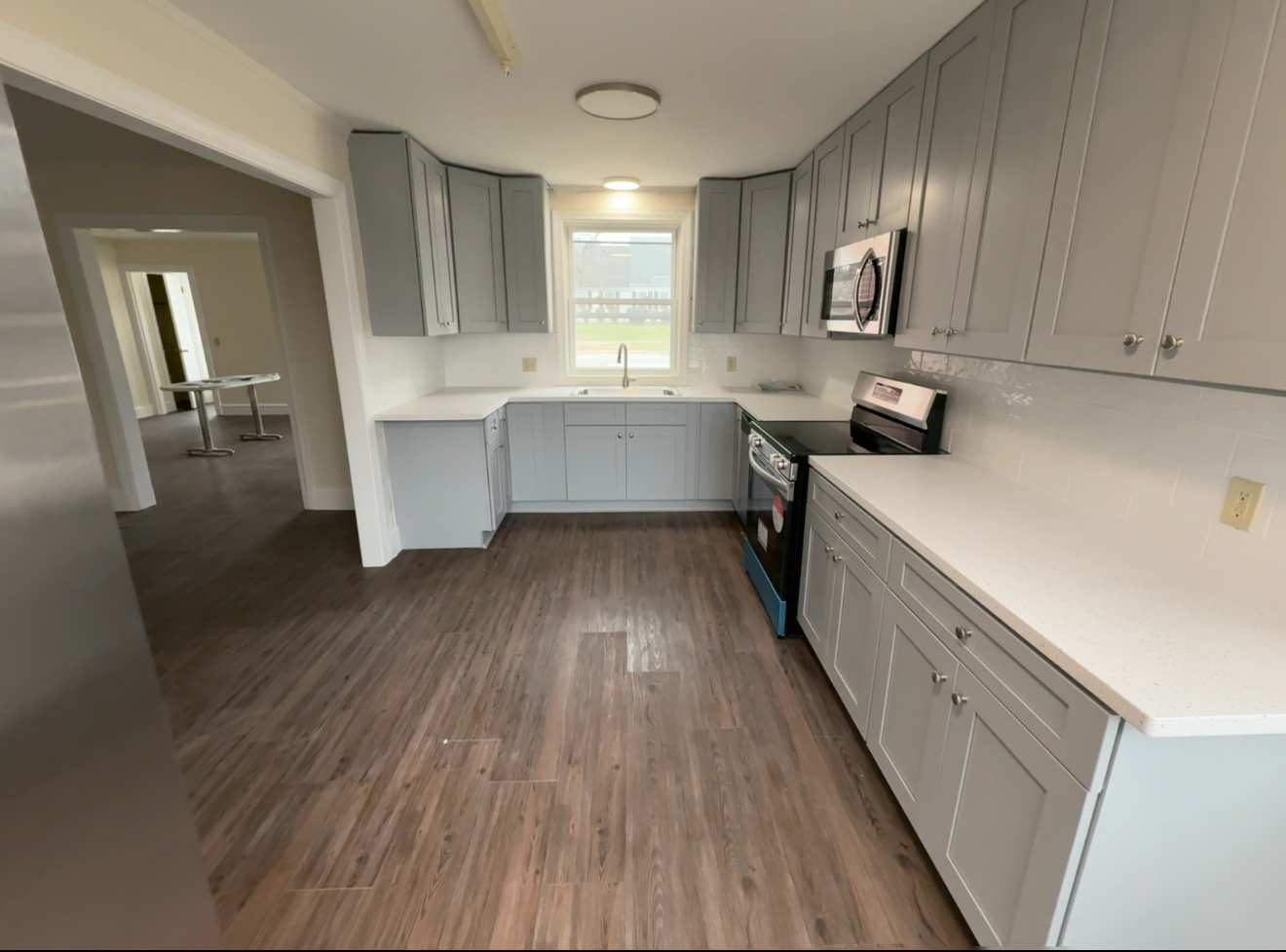 A modern kitchen with gray cabinets, white countertops, stainless steel appliances, and wood flooring. There is a window above the sink and an open doorway leading to another room.
