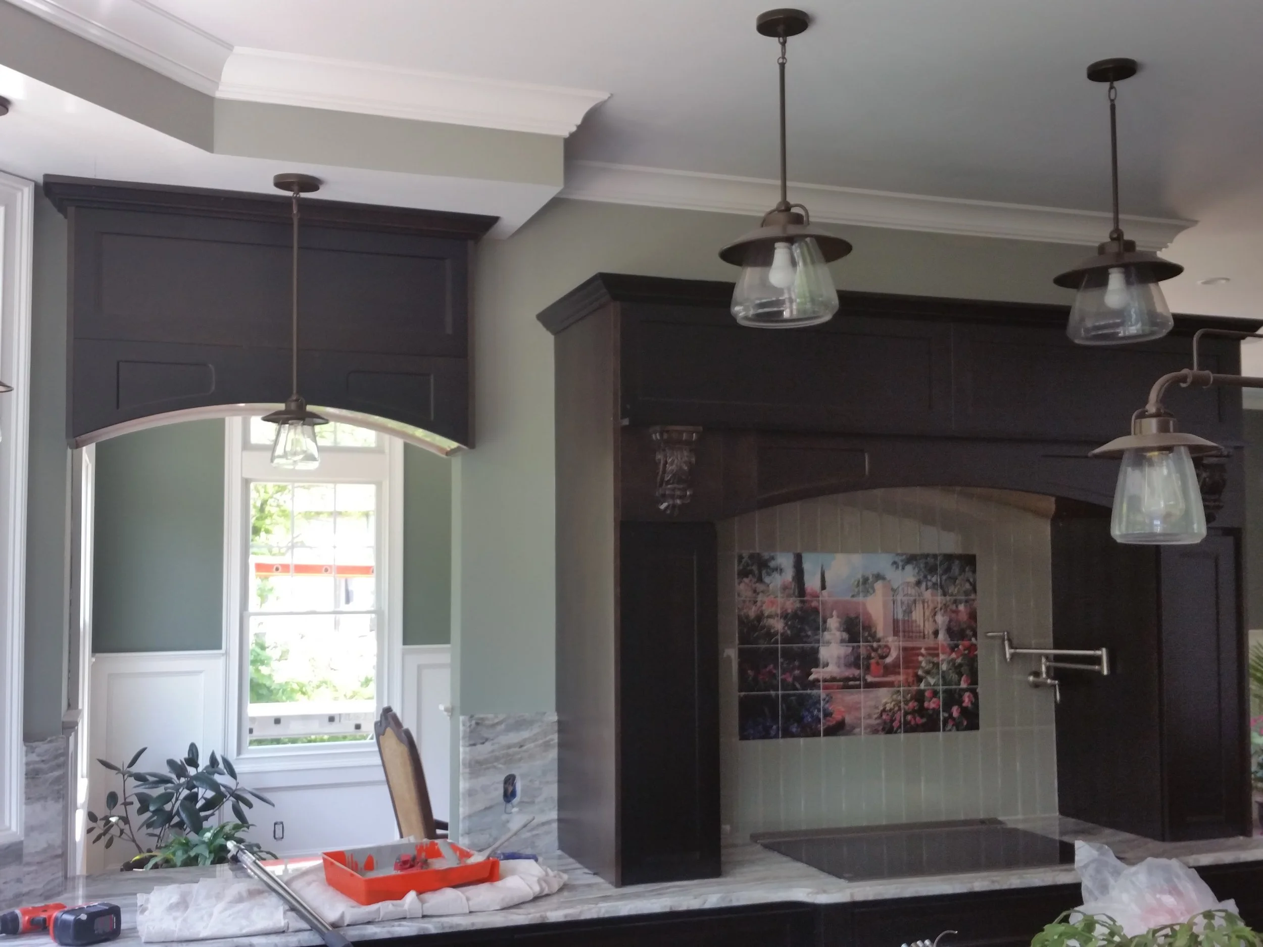 Interior view of a kitchen with black cabinetry, a window with a view outside, three hanging pendant lights with glass shades, a marble countertop, and a decorative tile mural of a fountain and garden scene behind the stove area.