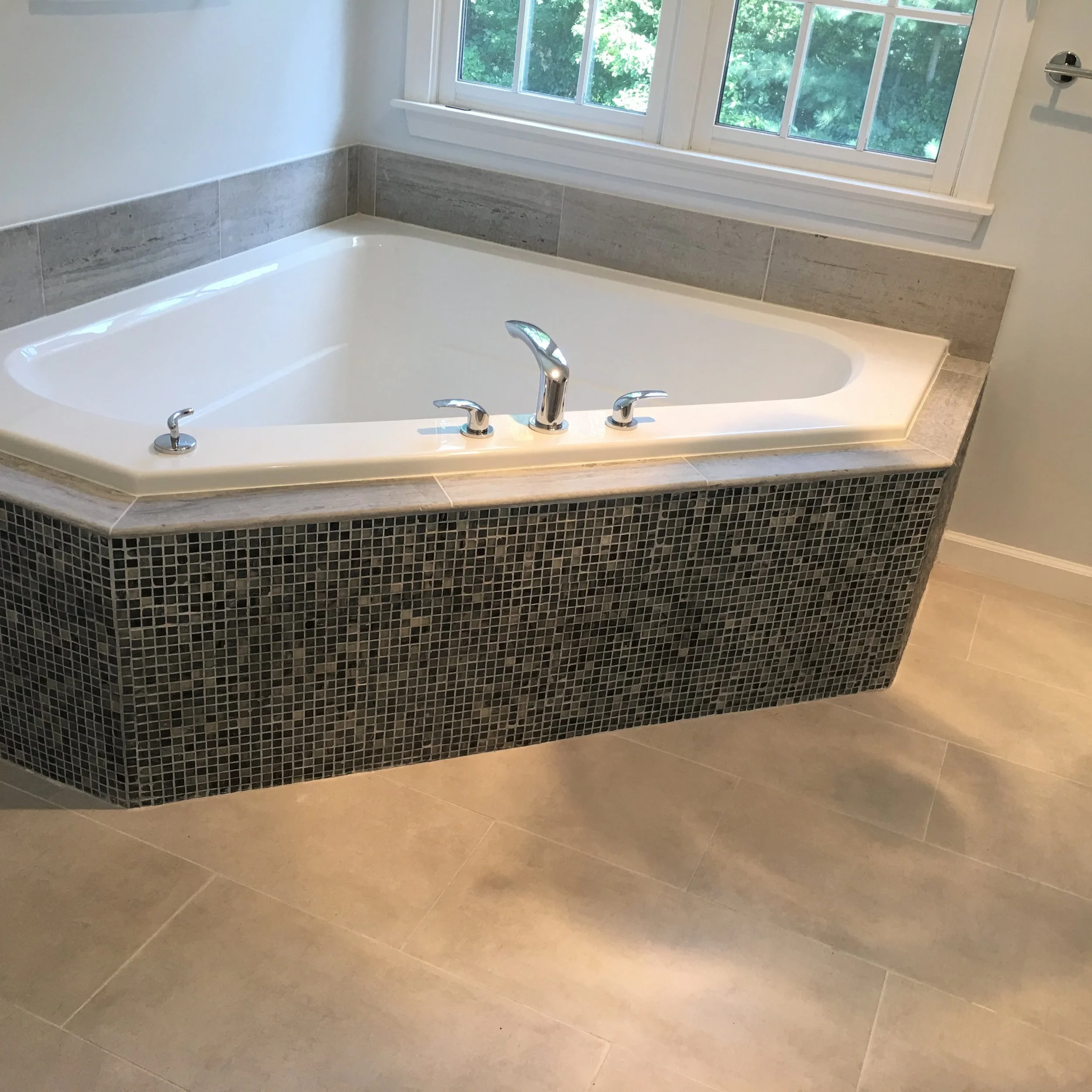 A built-in corner bathtub with a white interior, silver faucet, and controls, surrounded by gray tiles, positioned next to a large window with a view of green trees outside.