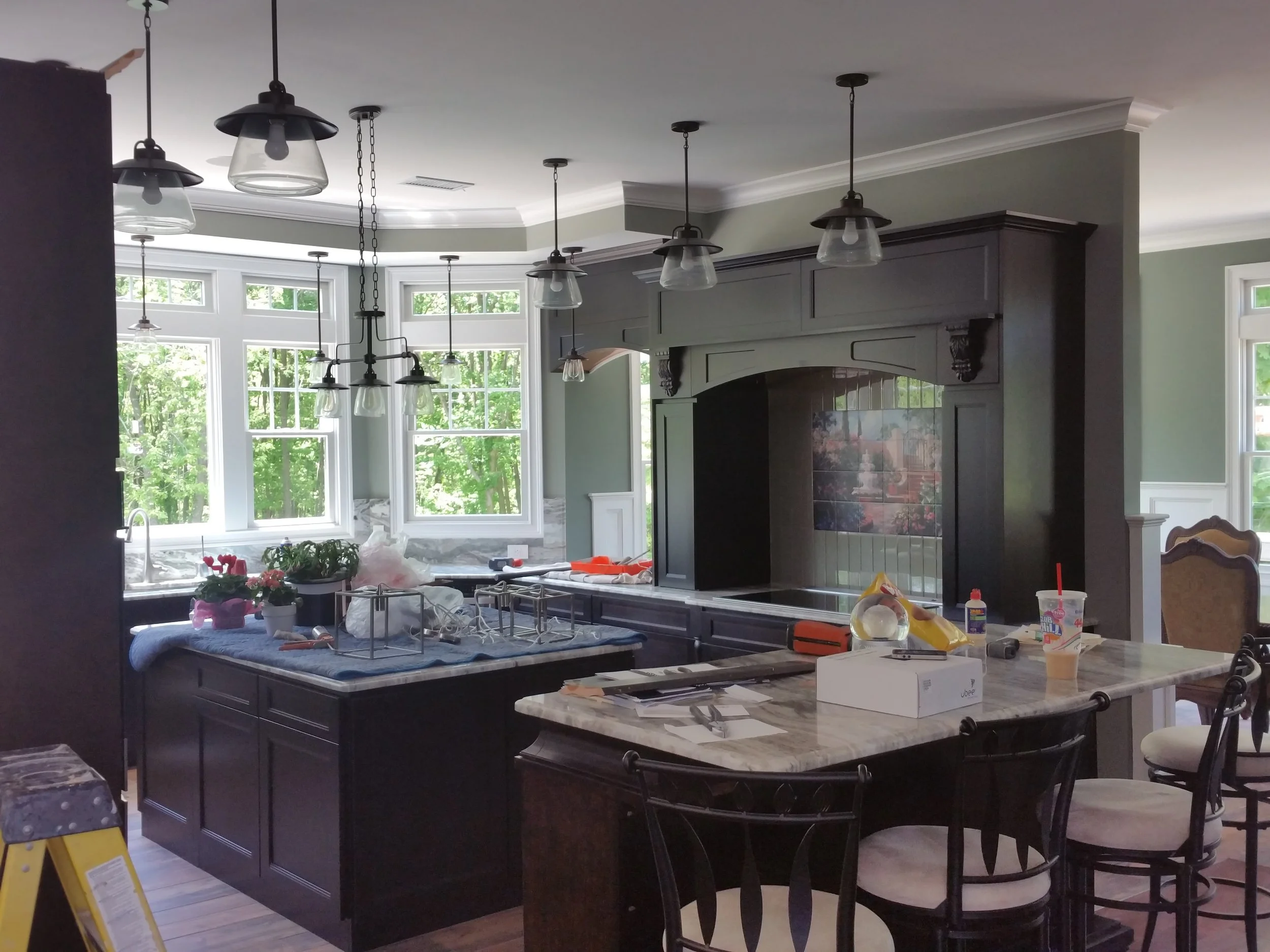 Kitchen renovation in progress with dark cabinetry, hanging light fixtures, and windows overlooking a wooded area.