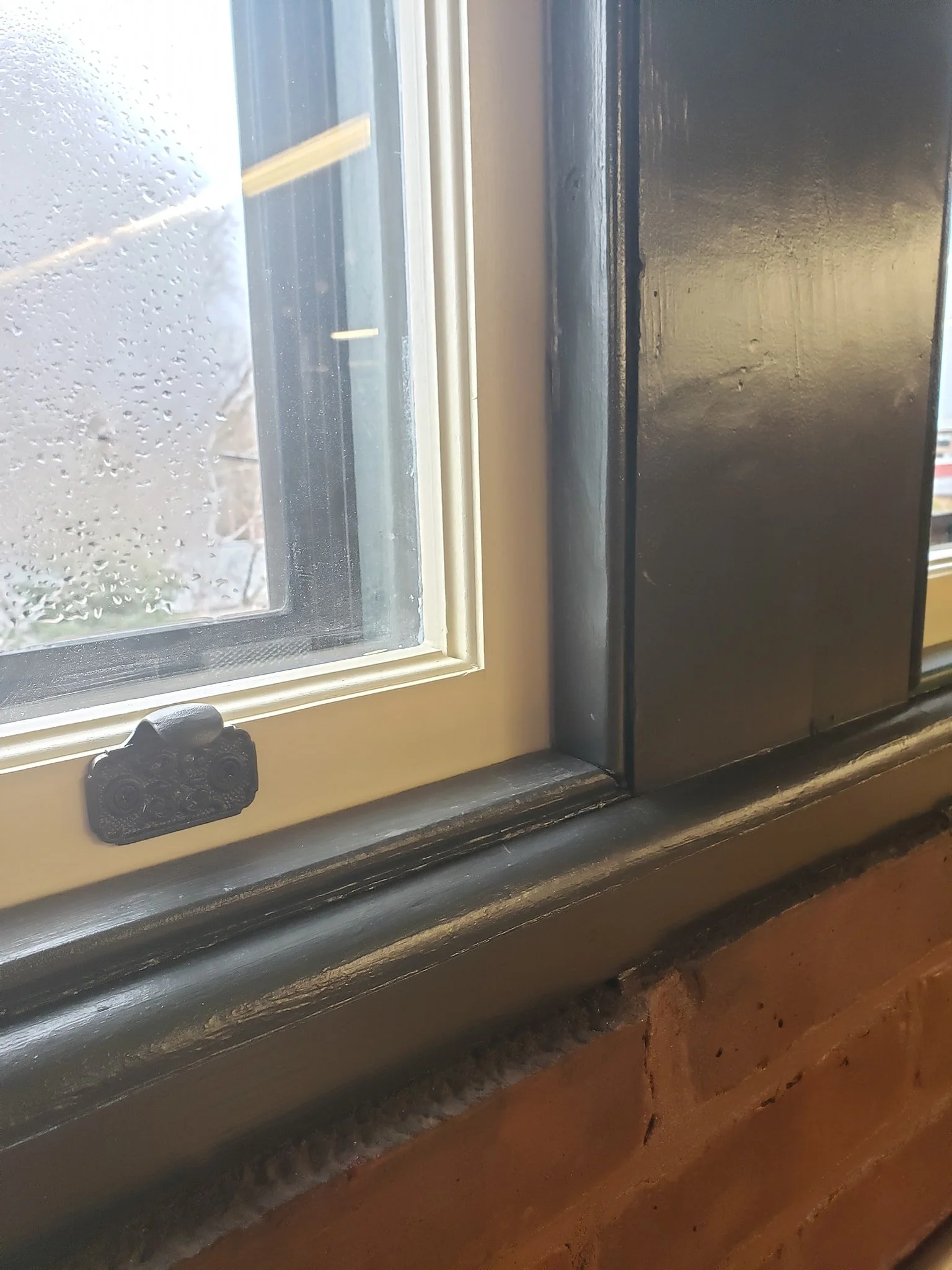 Close-up of a window with a white frame, black latch, and wet window glass, with raindrops outside and a brick wall below.