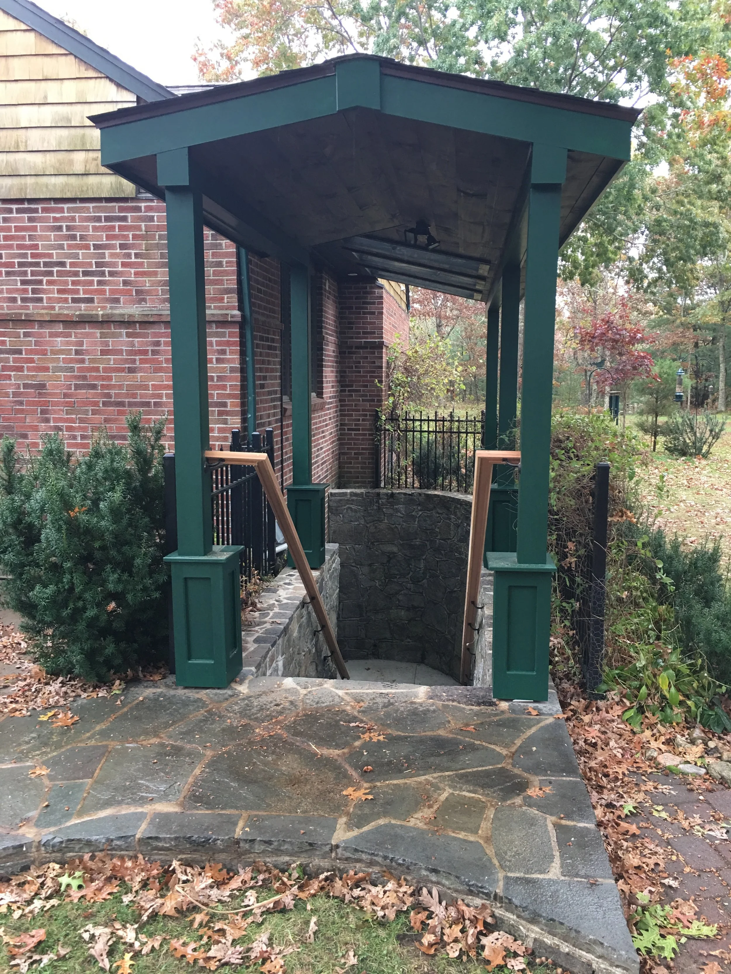 An outdoor entrance with a stone pathway leading down to a small set of stairs. The stairs are enclosed by a brick wall and metal fencing. A wooden railing is still being installed on one side, leaning against the green columns supporting a small roof in front of a brick house surrounded by trees with fall foliage.