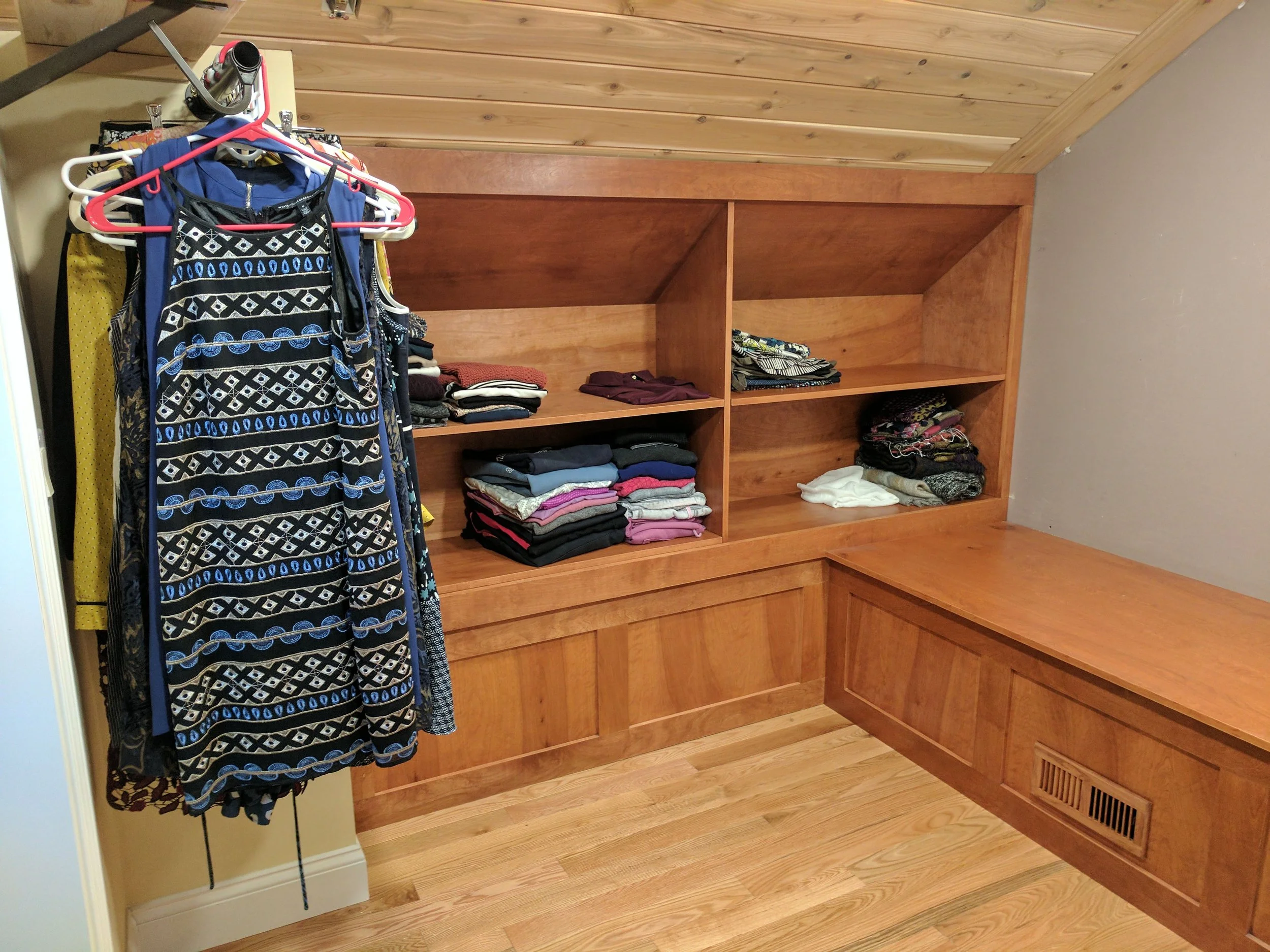Wooden built-in shelves with folded clothes and an area for hanging garments. A clothing rack holds dresses and garments on hangers to the left of the shelves.