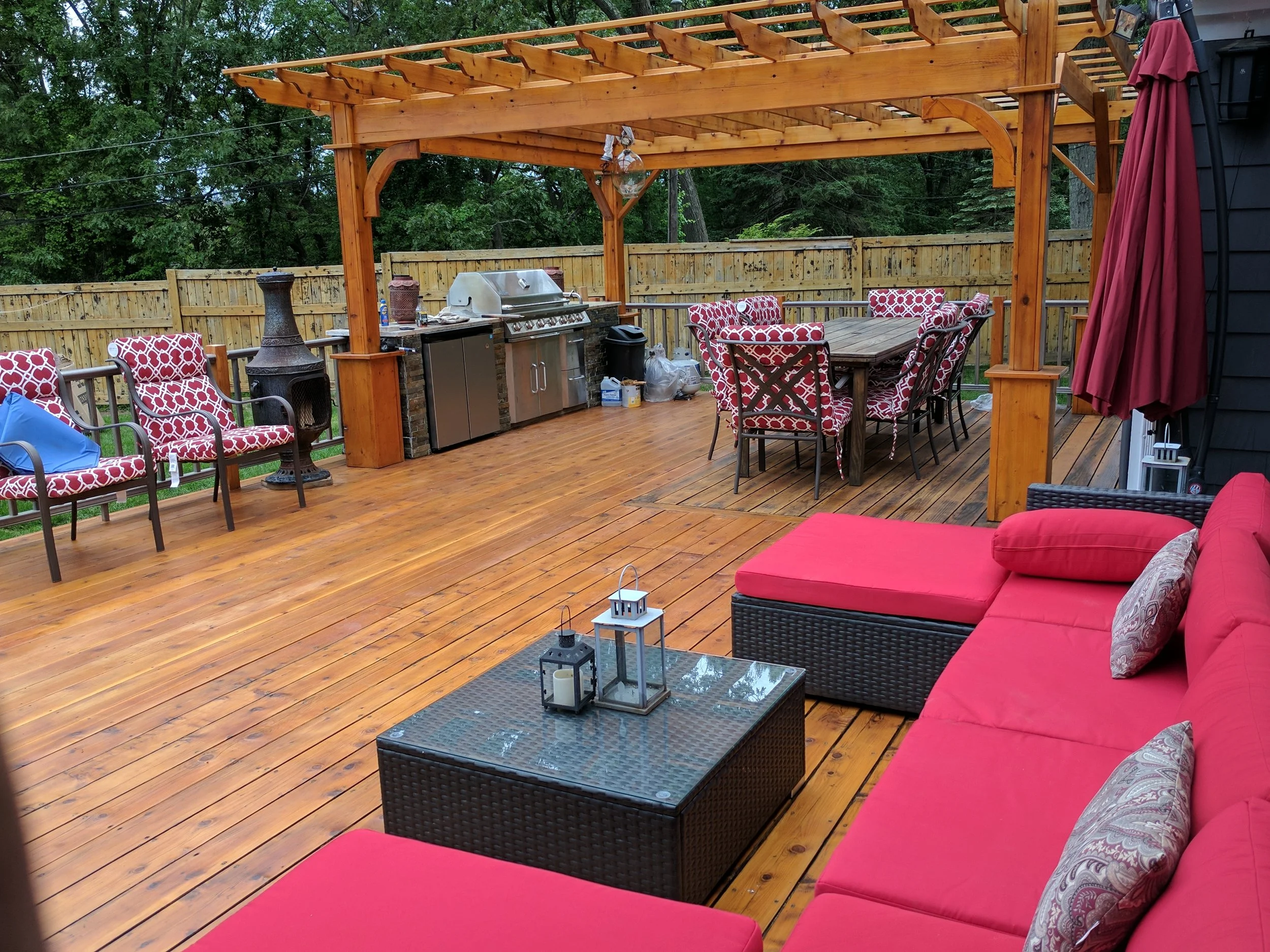 A wooden outdoor patio with red cushioned seating, a table with lanterns, a dining table with chairs, a grill, and a fence, surrounded by trees.