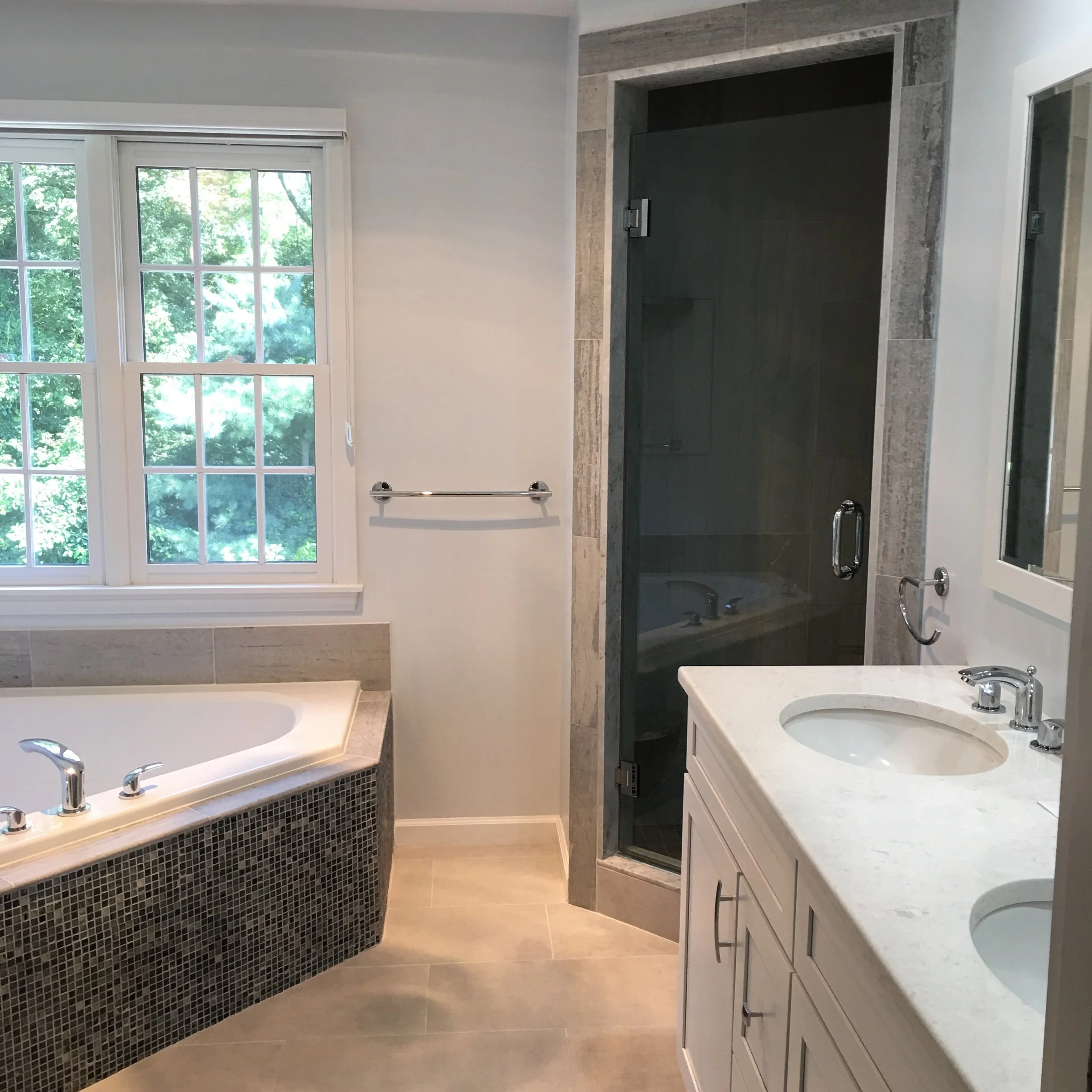 A bathroom featuring a large window over a bathtub with a mosaic tile exterior, a glass shower enclosure, and a white vanity with two sinks.