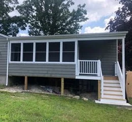 A gray manufactured home with a white porch and stairs, elevated on wooden supports, surrounded by green grass and trees.