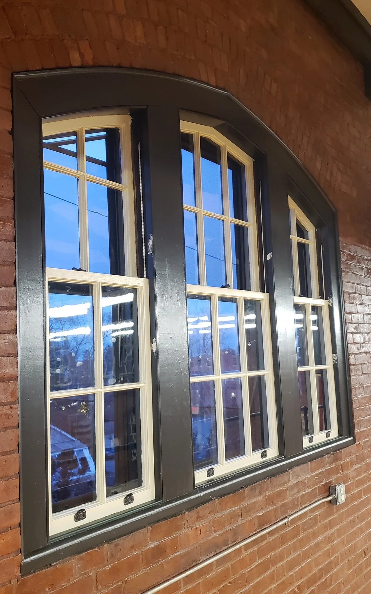 A large, decorative window with white divided panes and black trim, set into a red brick wall with an arched top, and an electrical conduit running along the wall below.