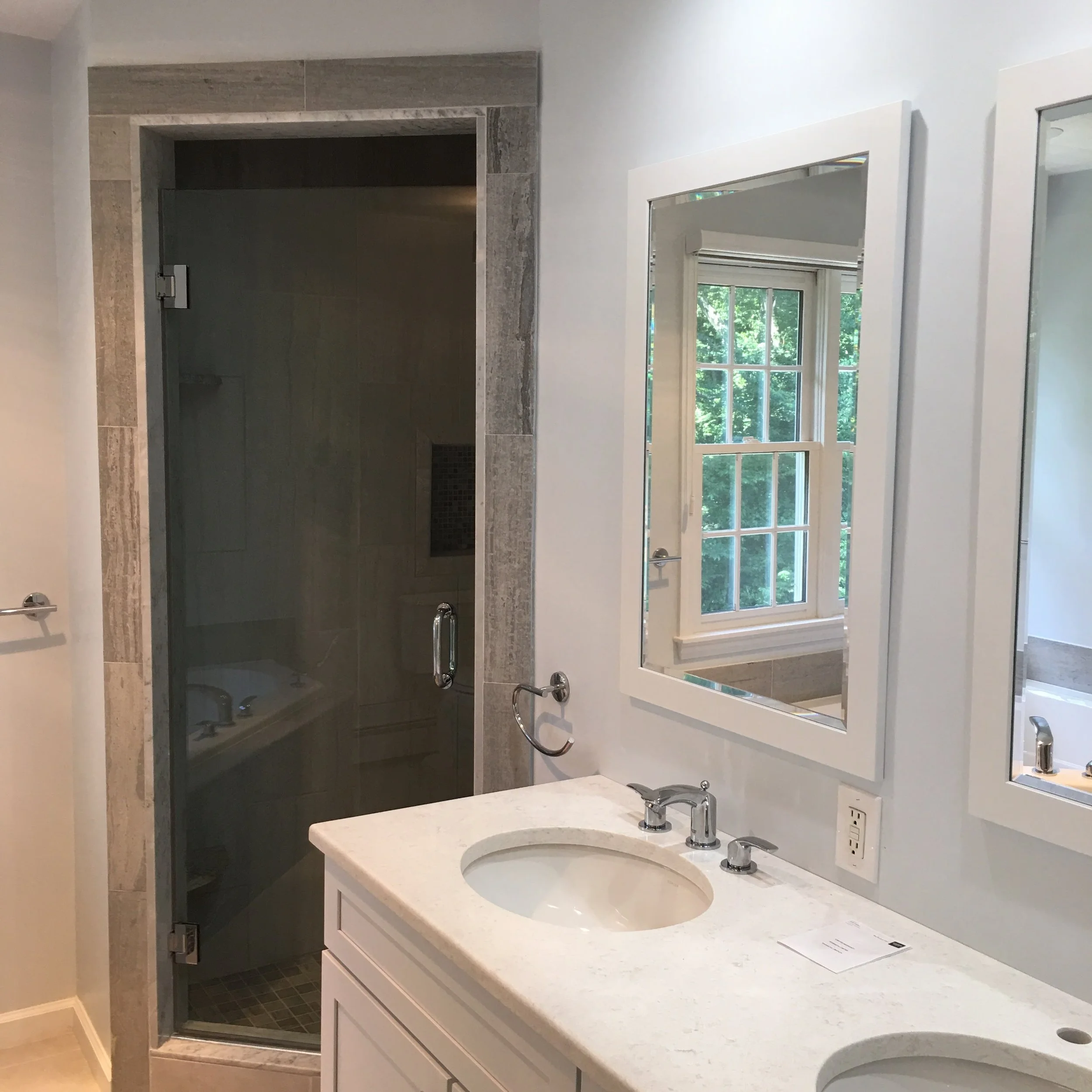 Bathroom with a white vanity, two sinks, and mirrors, next to a shower with a glass door and a window showing green trees outside.