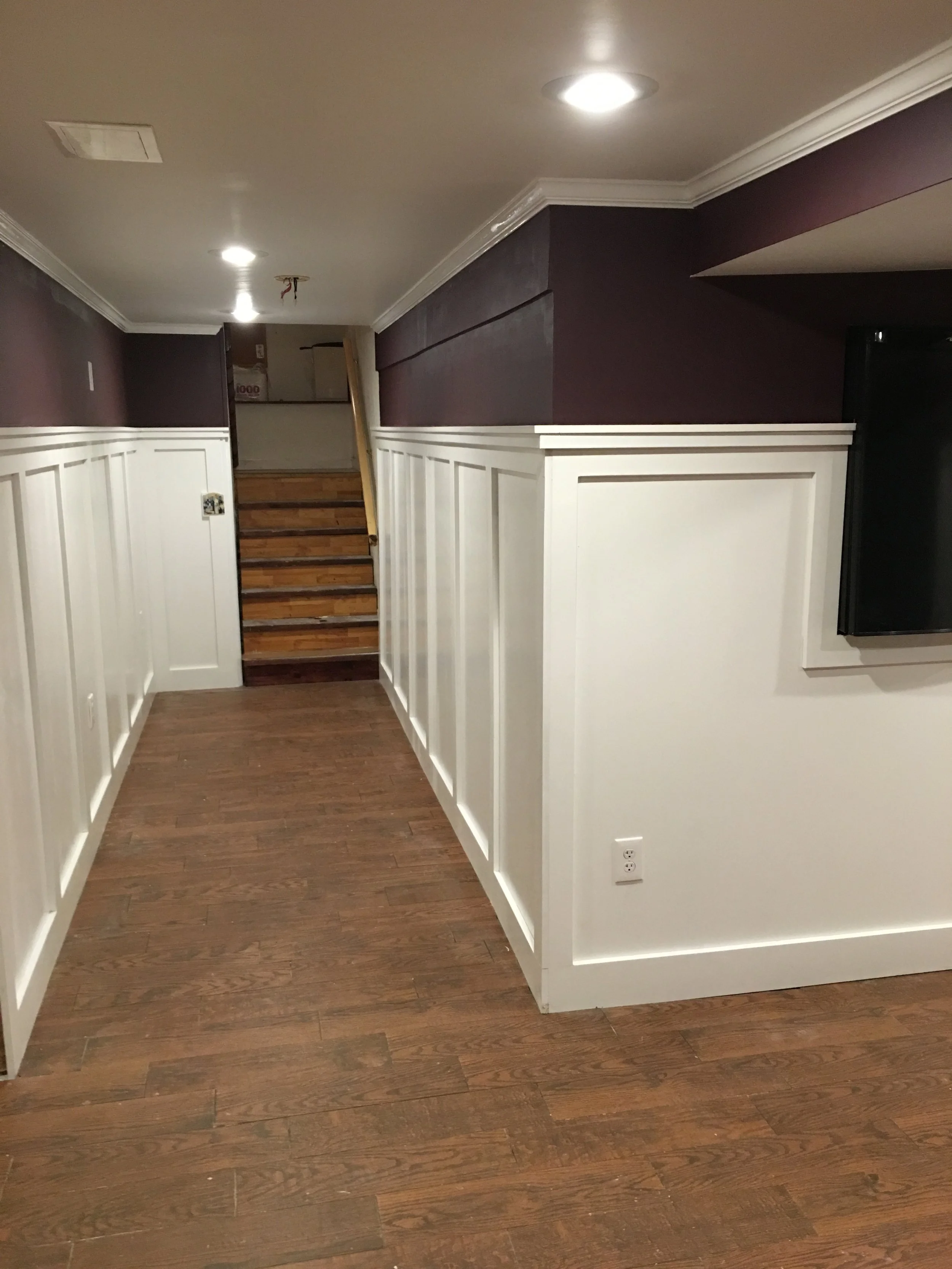 Interior view of a basement or lower-level room showing a short staircase with wooden steps leading up. The floor is wooden, and the walls are decorated with white wainscoting and dark purple paint above. Recessed ceiling lights and an electrical out