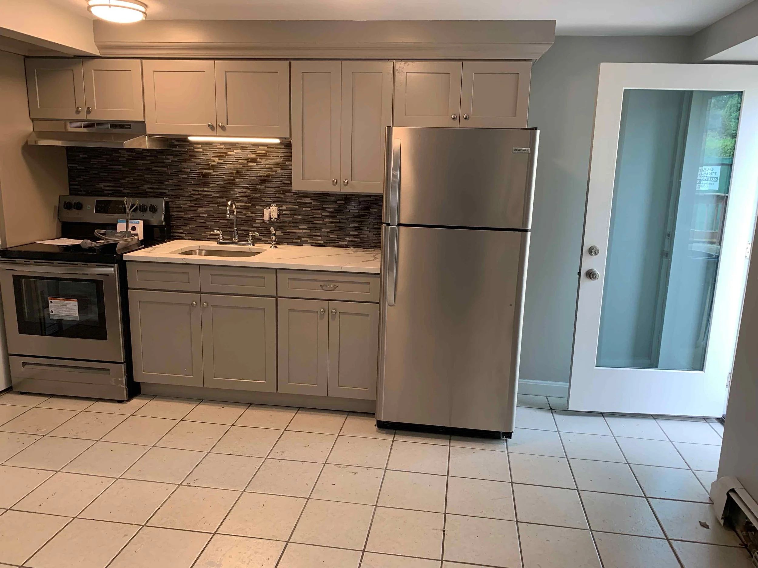 Kitchen with beige cabinets, stainless steel stove, refrigerator, and a mosaic tile backsplash.