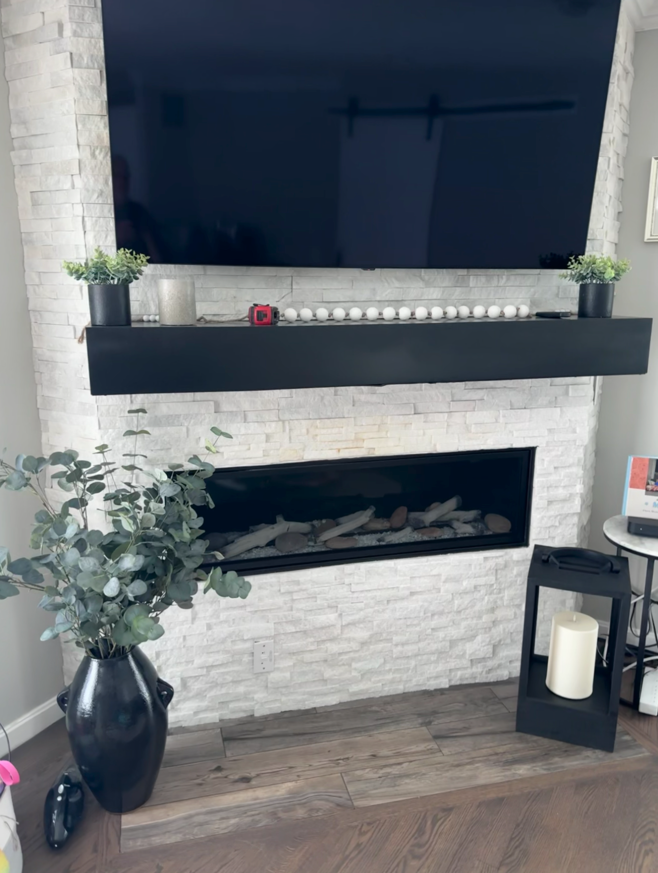 Living room fireplace with a mounted TV above a black mantel, decorated with plants, candles, and a red tape measure. There is a large potted plant on the floor and a candle holder next to a small table with a device and magazine.