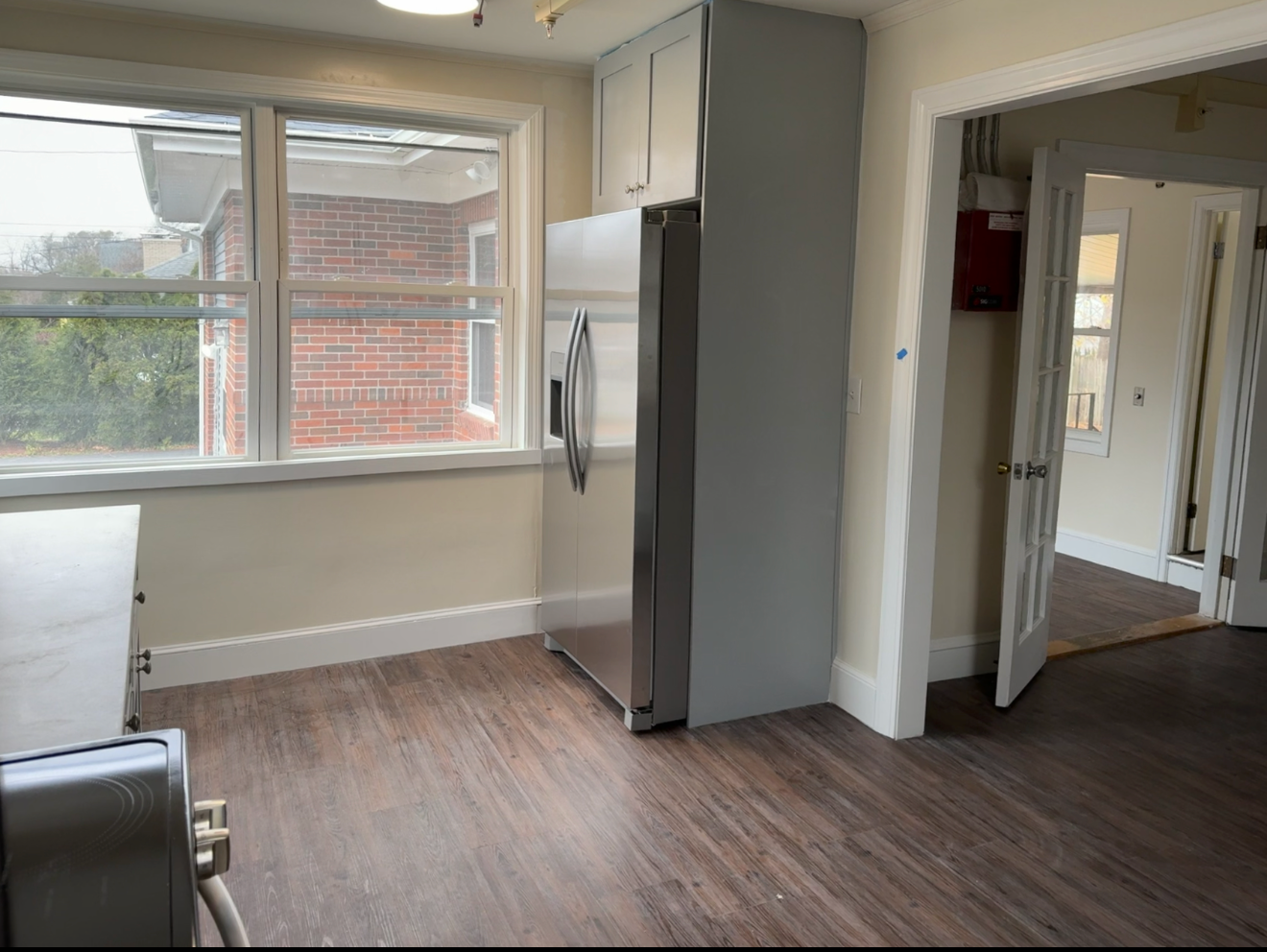 Empty kitchen with a large window, stainless steel refrigerator, and a doorway to another room with a fire alarm system on the wall.