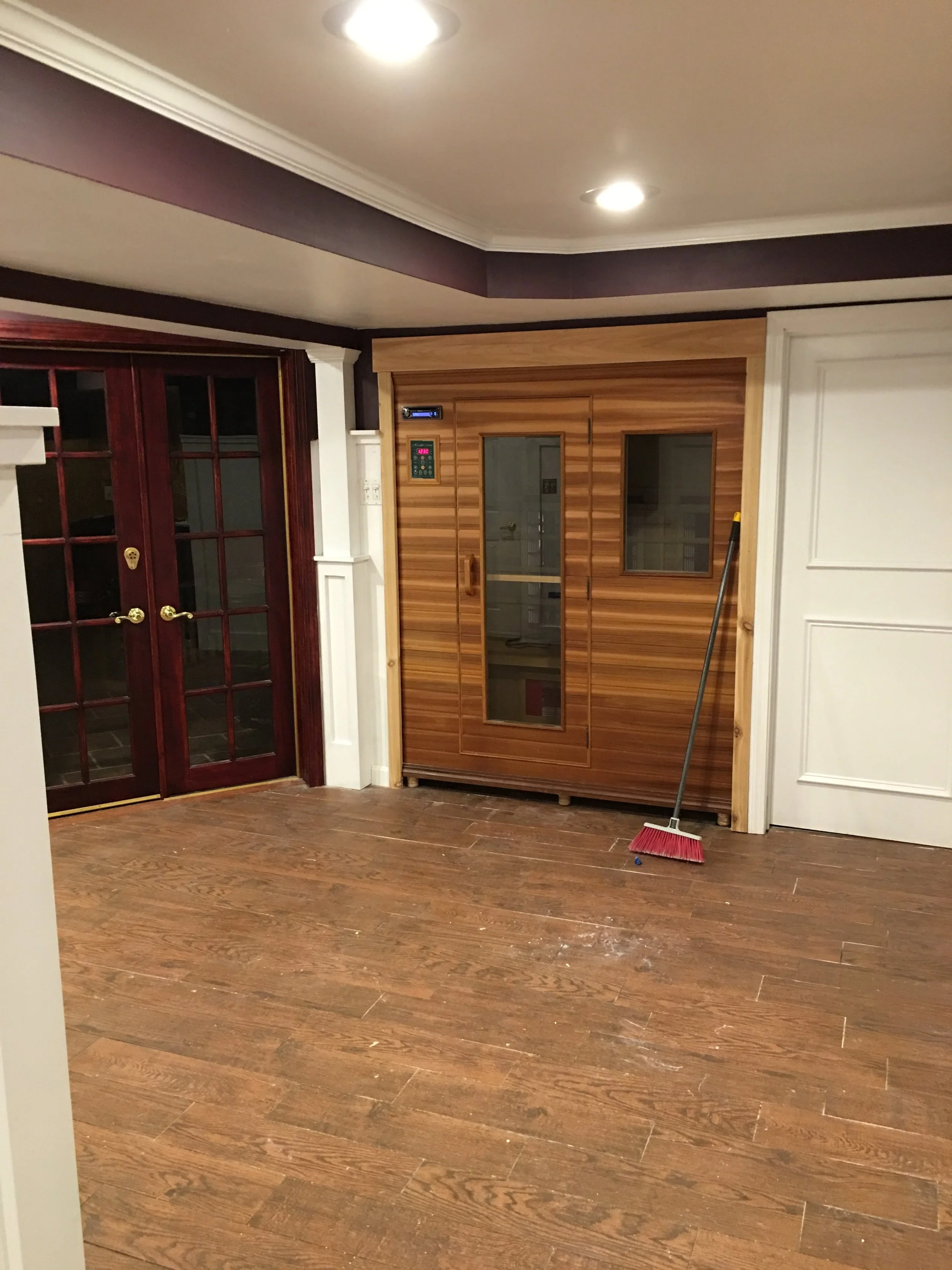 Living room with wooden flooring, French doors, and a sauna cabinet. A broom leans against the sauna. Recessed ceiling lights are on.