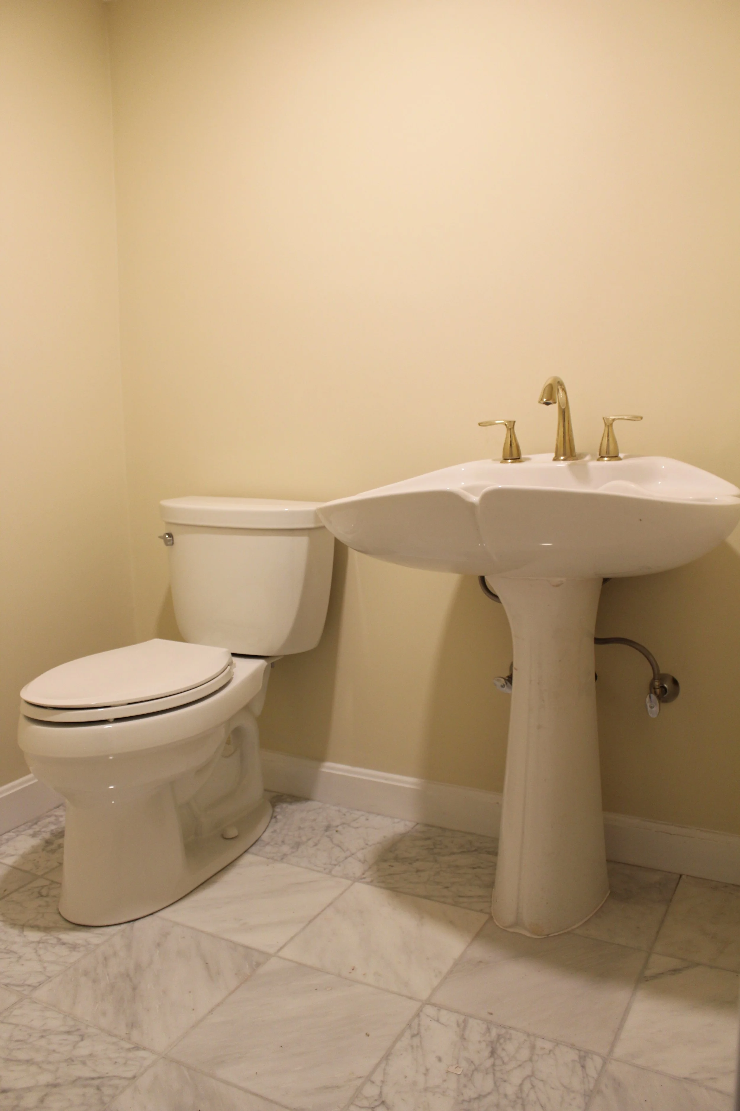 Bathroom with a white toilet next to a white pedestal sink with gold fixtures, on a marble floor, against a beige wall.