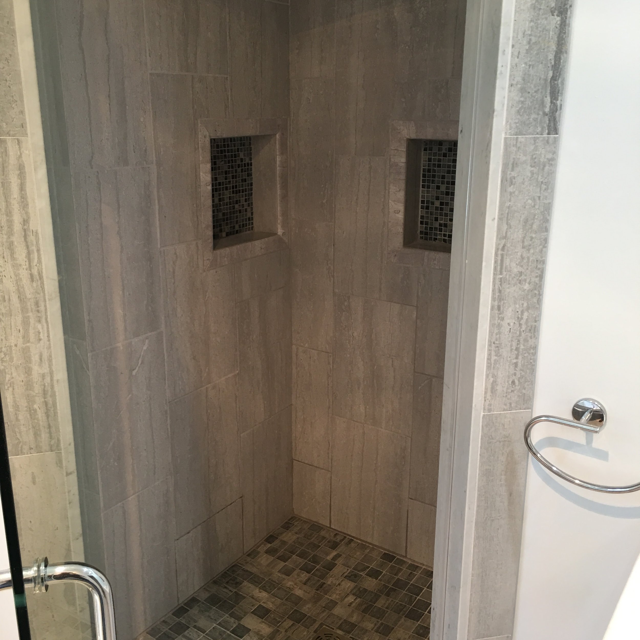 A modern walk-in shower with beige tiles on the walls and small mosaic tiles in shades of brown and beige on the floor. There are two built-in recessed shelves with dark mosaic tiling.