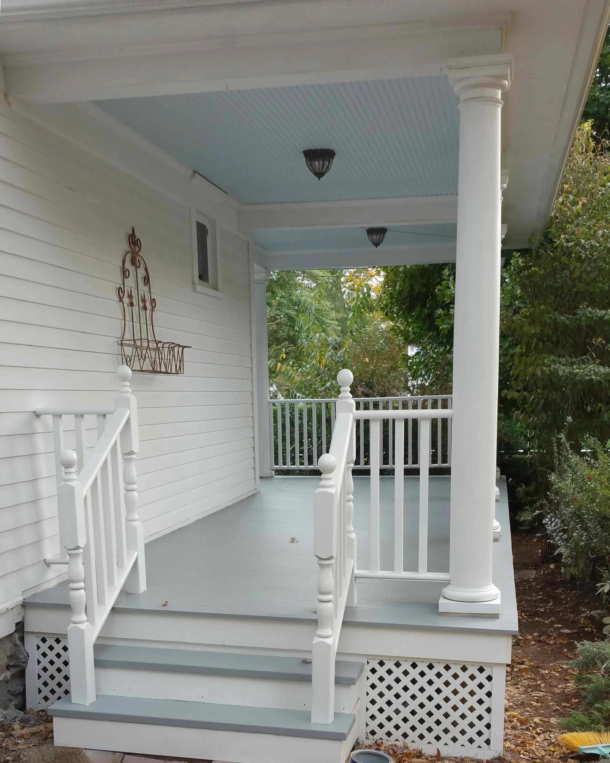 Front porch of a house with white railings, steps, and column, and a light blue ceiling. Green trees in the background.