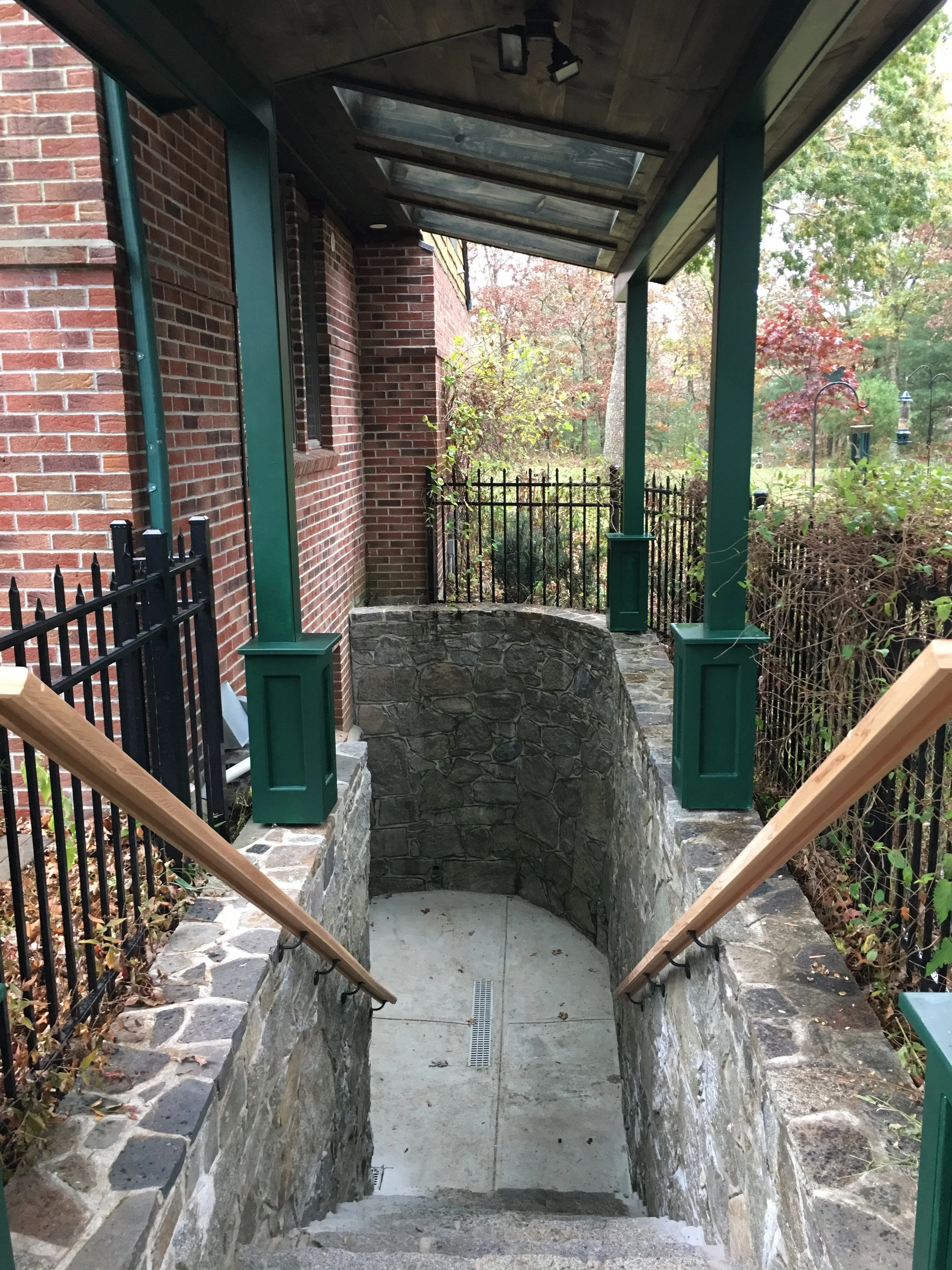 An outdoor staircase leading down to a basement or cellar, with stone walls, concrete steps, and wooden handrails, adjacent to a brick building with green trim and a glass-enclosed roof.