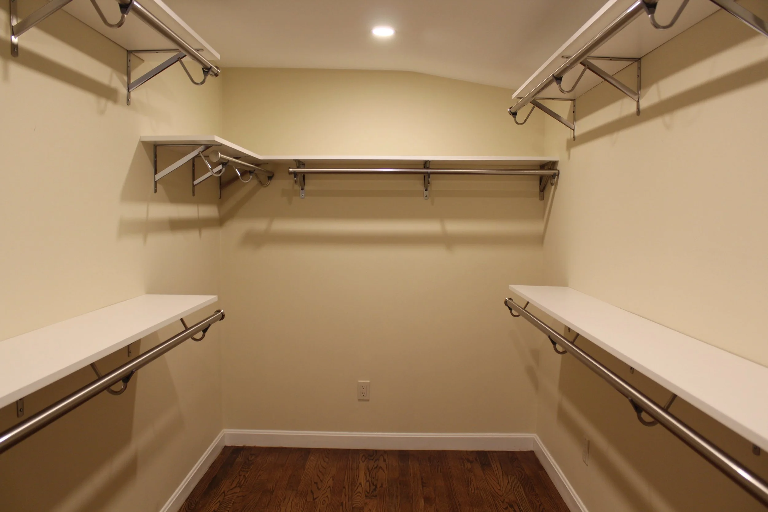 Empty walk-in closet with beige walls, wooden floor, and white shelving with metal rods for hanging clothes.