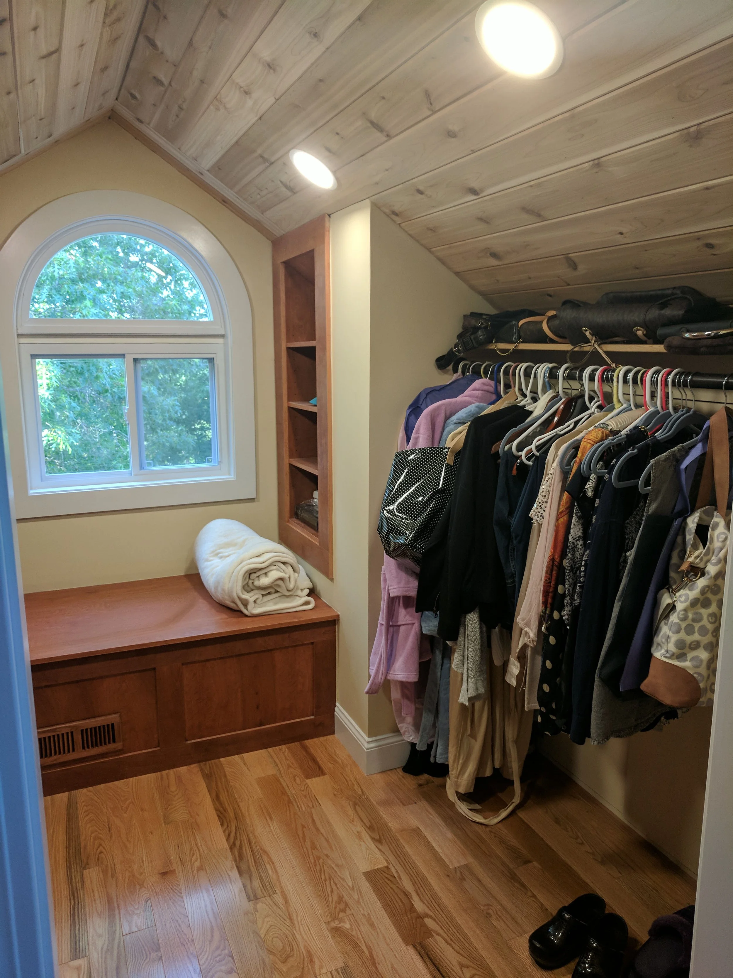 Small walk-in closet with a window, wooden ceiling, and a bench with a folded blanket. Closet has hanging clothes, shelves, and shoes.
