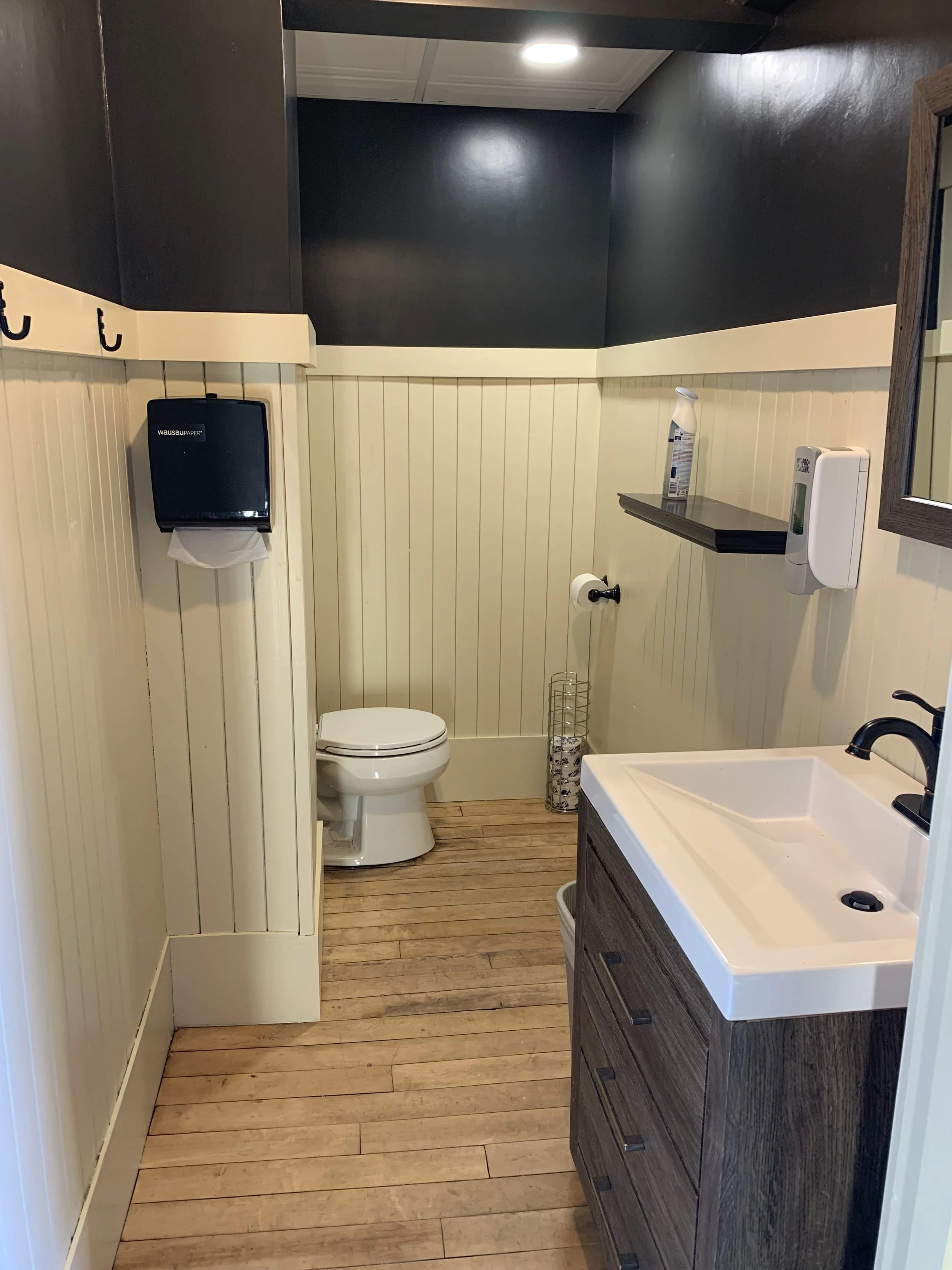Small bathroom with wood floor and beige paneled walls, containing a toilet, a sink with a dark wood cabinet, a mirror, a soap dispenser, a hand sanitizer, a shelf with bathroom items, a toilet paper holder, a wastebasket, and a wall-mounted paper towel dispenser.