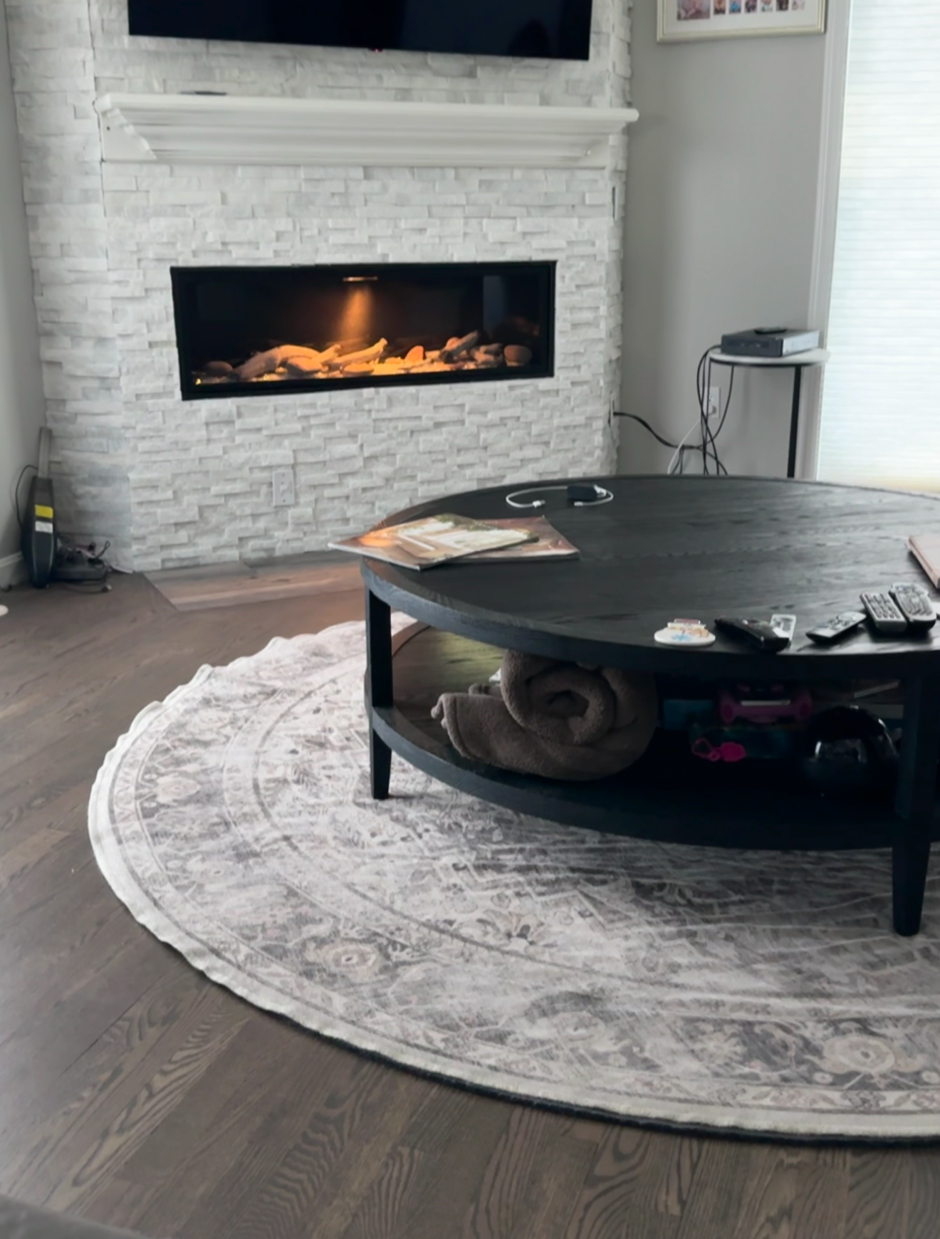 Living room with a white stone fireplace, a flat-screen TV mounted above, a round black coffee table with remote controls, magazines, a folded towel underneath, and a patterned rug on hardwood floors, with a corner table holding a cable box and wires.