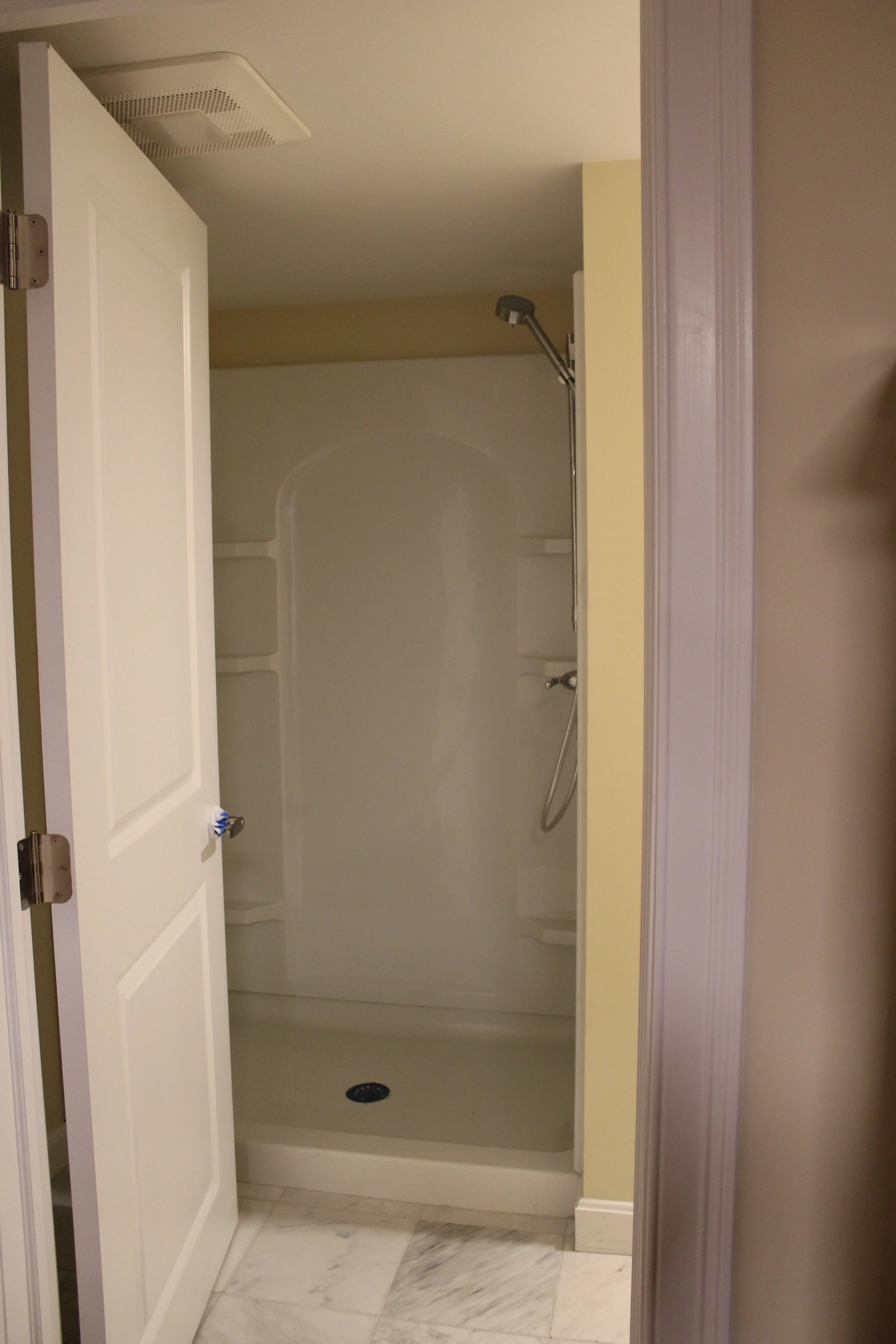 Empty shower stall with built-in shelves, a handheld showerhead, and a drain at the bottom, viewed through an open door in a bathroom with marble flooring.