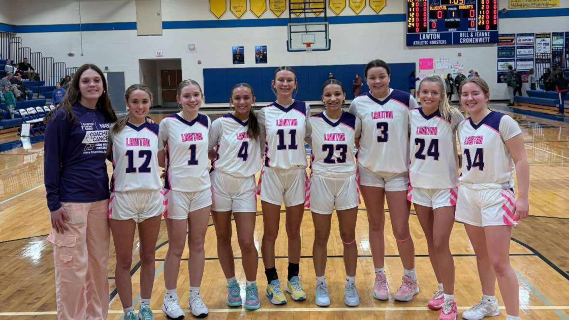 Group of nine young female volleyball players in team uniforms and one coach standing on a basketball court in a gymnasium.