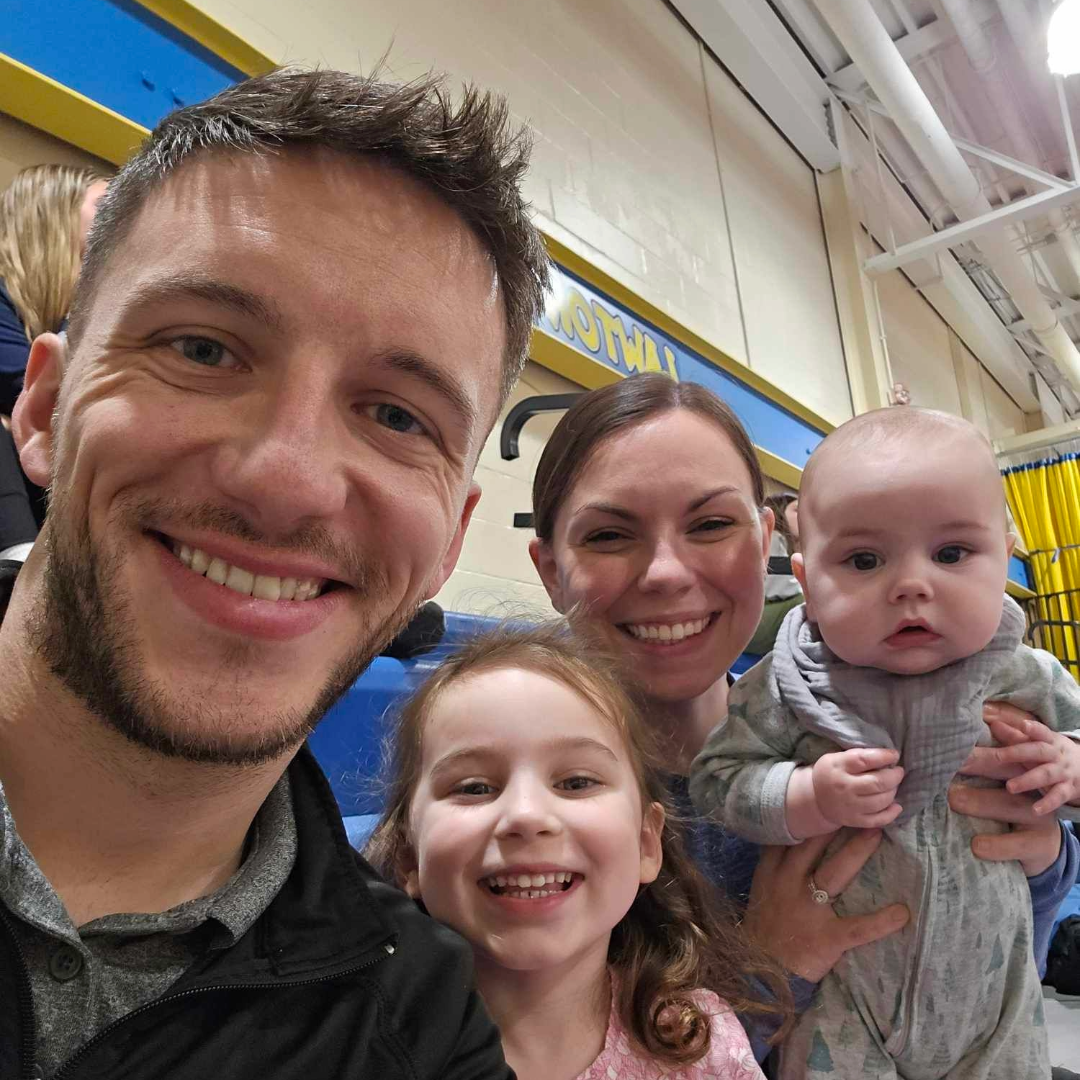 A family of four taking a selfie at an indoor ice skating rink. The father is on the left, smiling, with light brown hair and a beard. Next to him is a young girl with curly brown hair smiling. Behind them is a woman with brown hair also smiling, holding a baby with a bald head and wearing a gray hoodie, who looks curious.