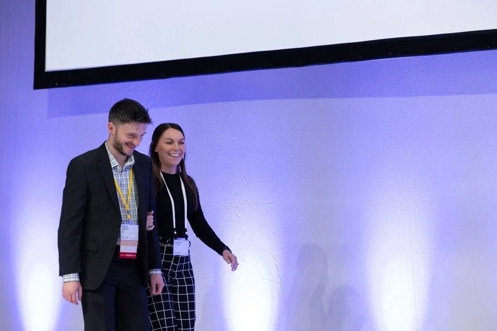 Two young adults, a man and a woman, walking and smiling in front of a light purple-blue gradient wall, possibly at a conference or event.
