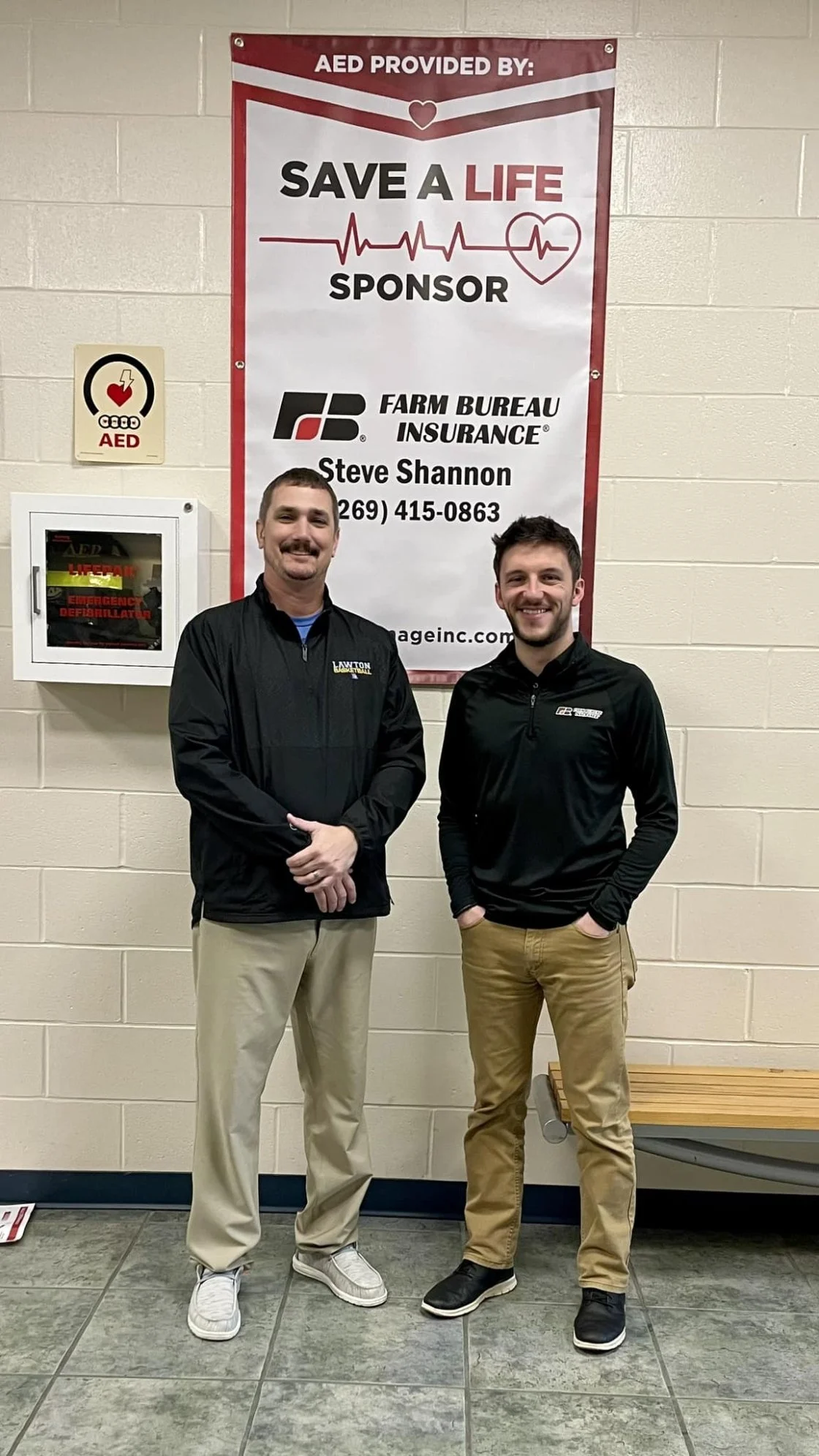 Two men standing in front of a large red and white sign that promotes AED donation for Life Save. The sign displays the sponsor's name, Steve Shannon's contact information, and the logo of Farm Bureau Insurance. The man on the left wears a black jacket with beige pants, and the man on the right wears a black jacket with tan pants. An AED cabinet is mounted on the wall to the left of the sign.