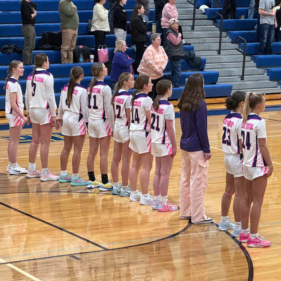 Women's basketball team standing in a line on a gymnasium court, wearing white uniforms with pink and purple accents, with spectators seated and standing in the background.