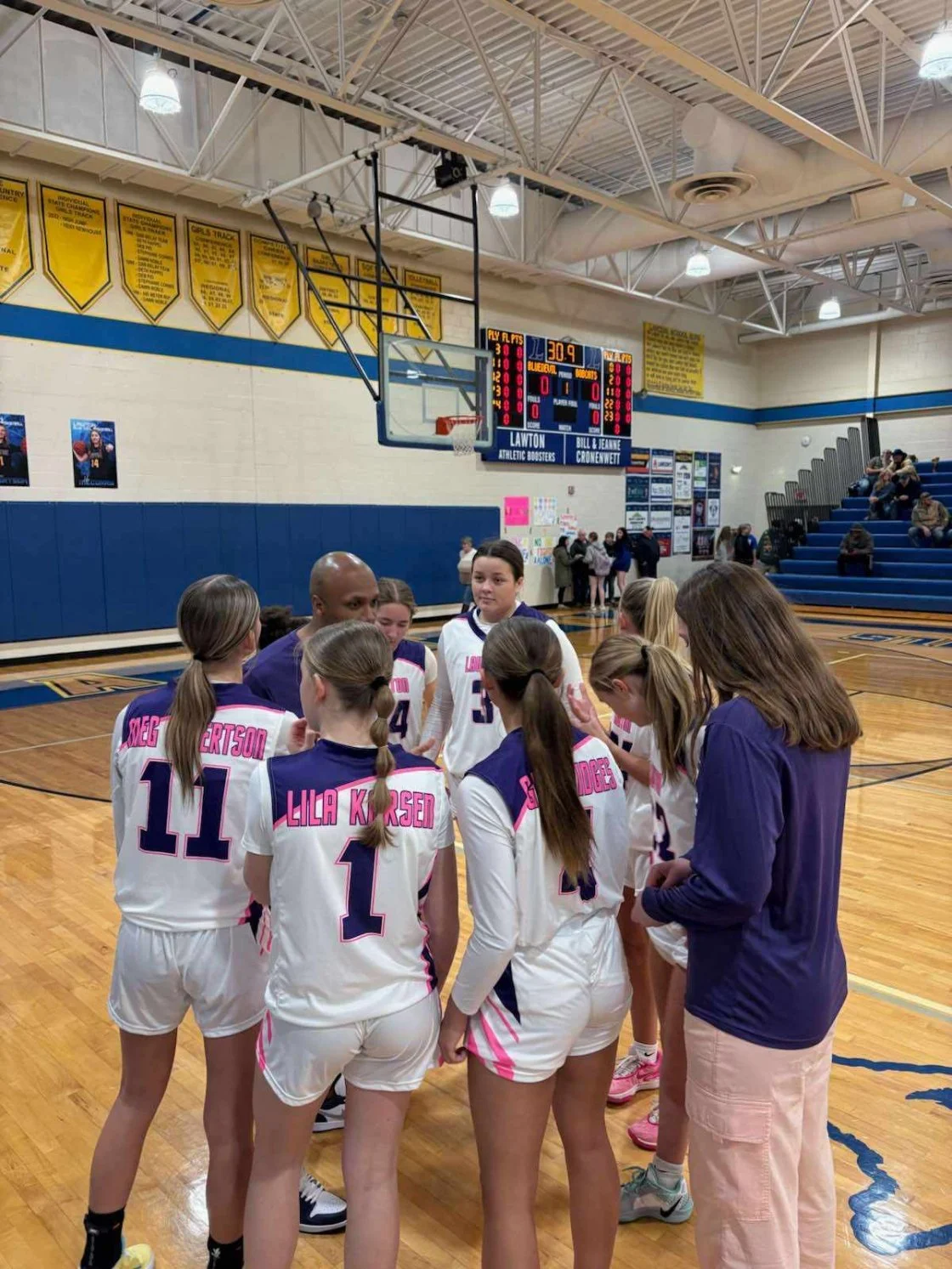 Girls' basketball team huddled in a gym, coach speaking to them, with scoreboard and bleachers in background.