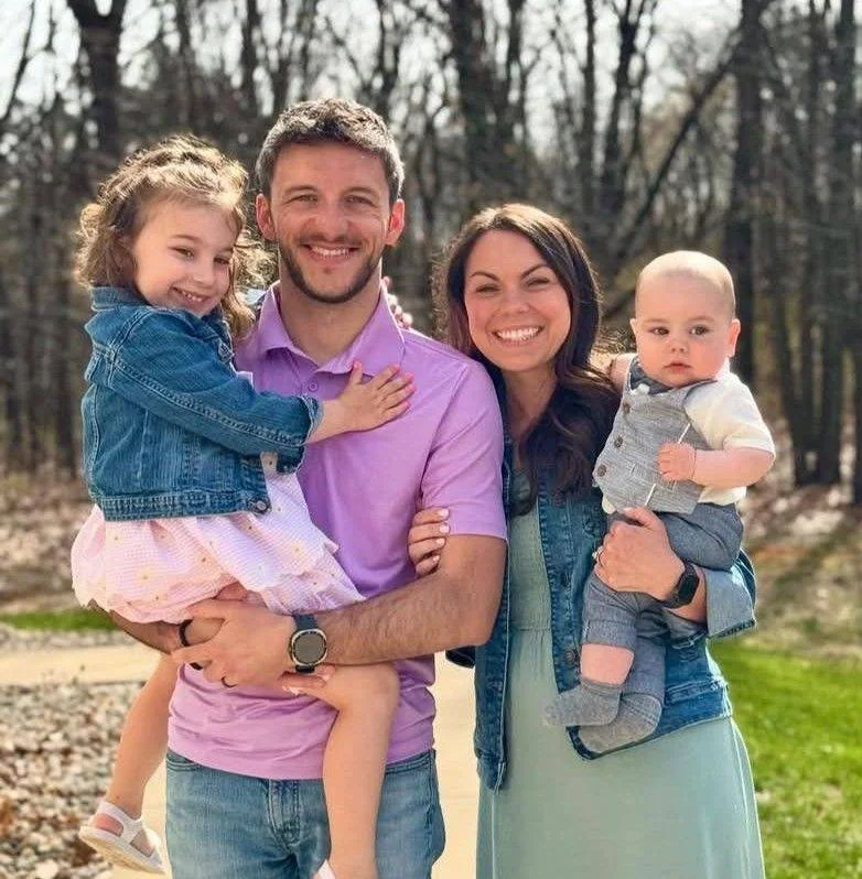 A smiling family of four outdoors in a wooded area during daytime, with the father holding a young girl and the mother holding a baby boy.