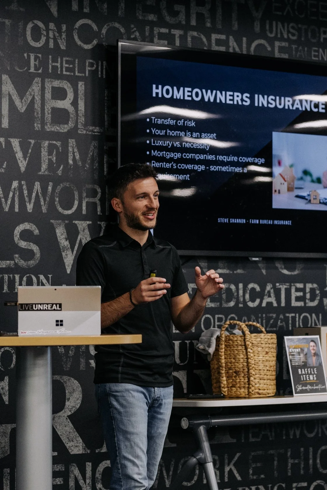Man giving a presentation about homeowners insurance in front of a large screen, with a laptop and basket on a table nearby.