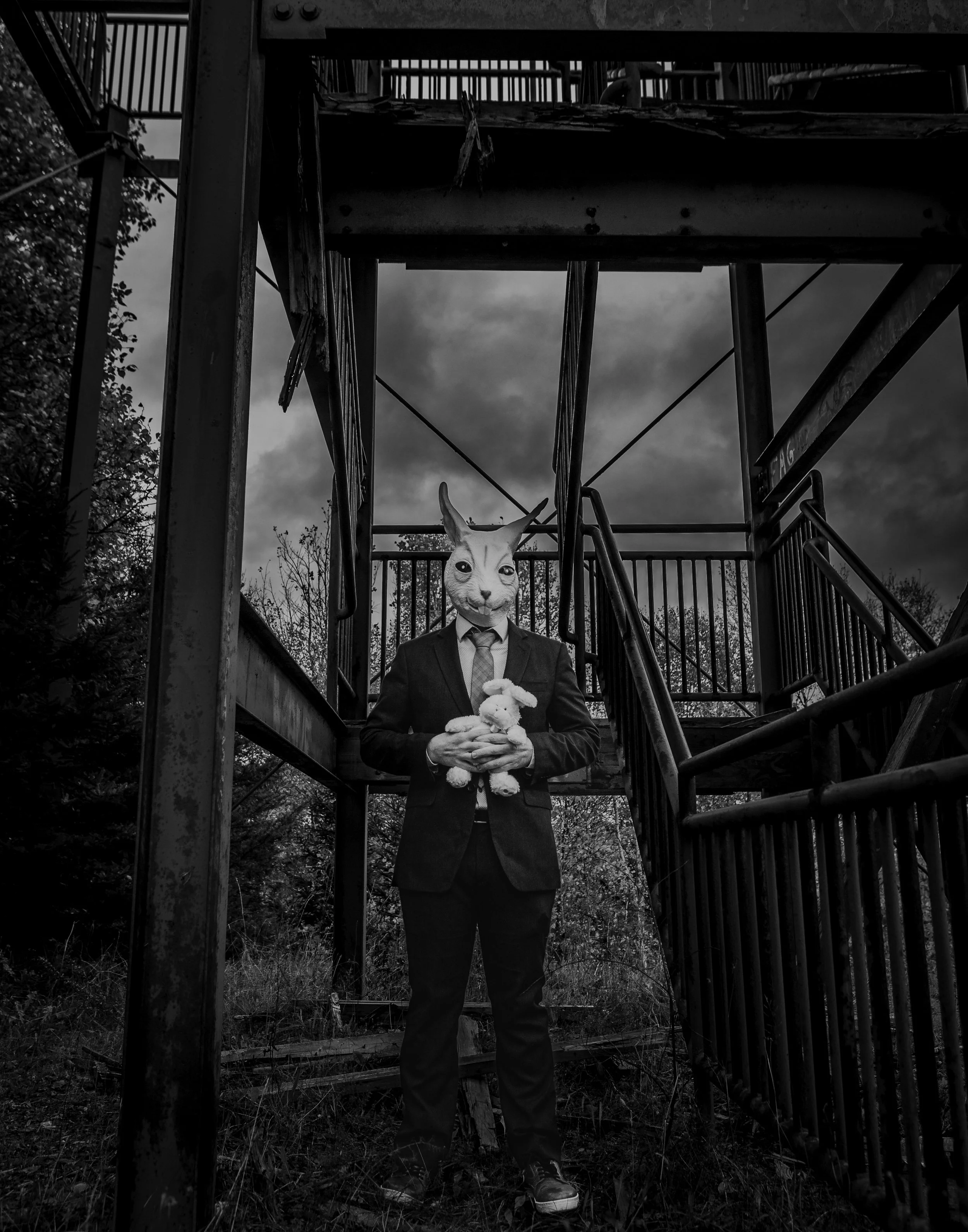 A person in a suit with a rabbit’s face stands on the ground, holding a stuffed bunny in front of a dilapidated outdoor staircase with dark clouds overhead.