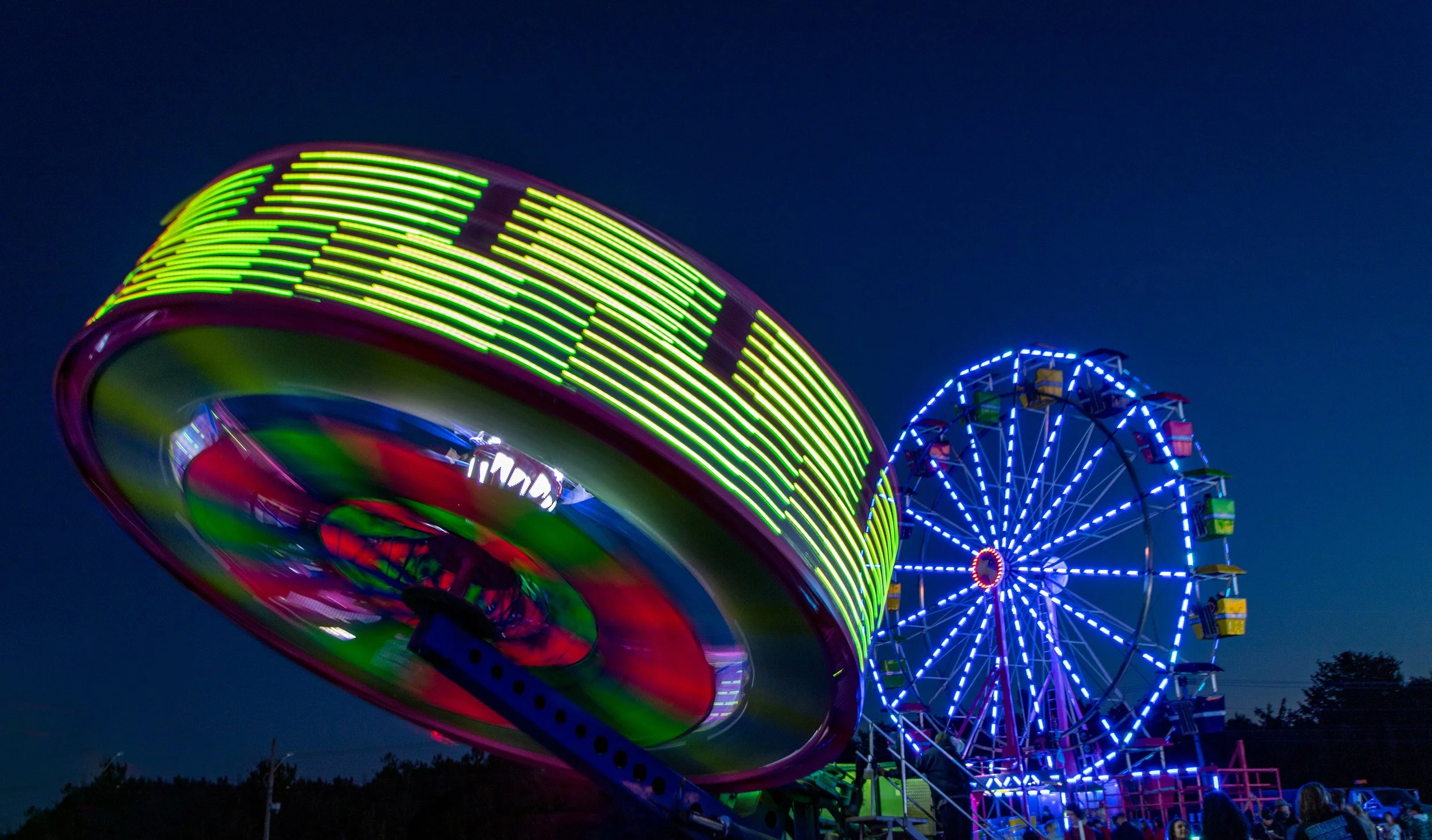 Colorful amusement park rides with neon lights at night, featuring a spinning ride in the foreground and a Ferris wheel in the background.