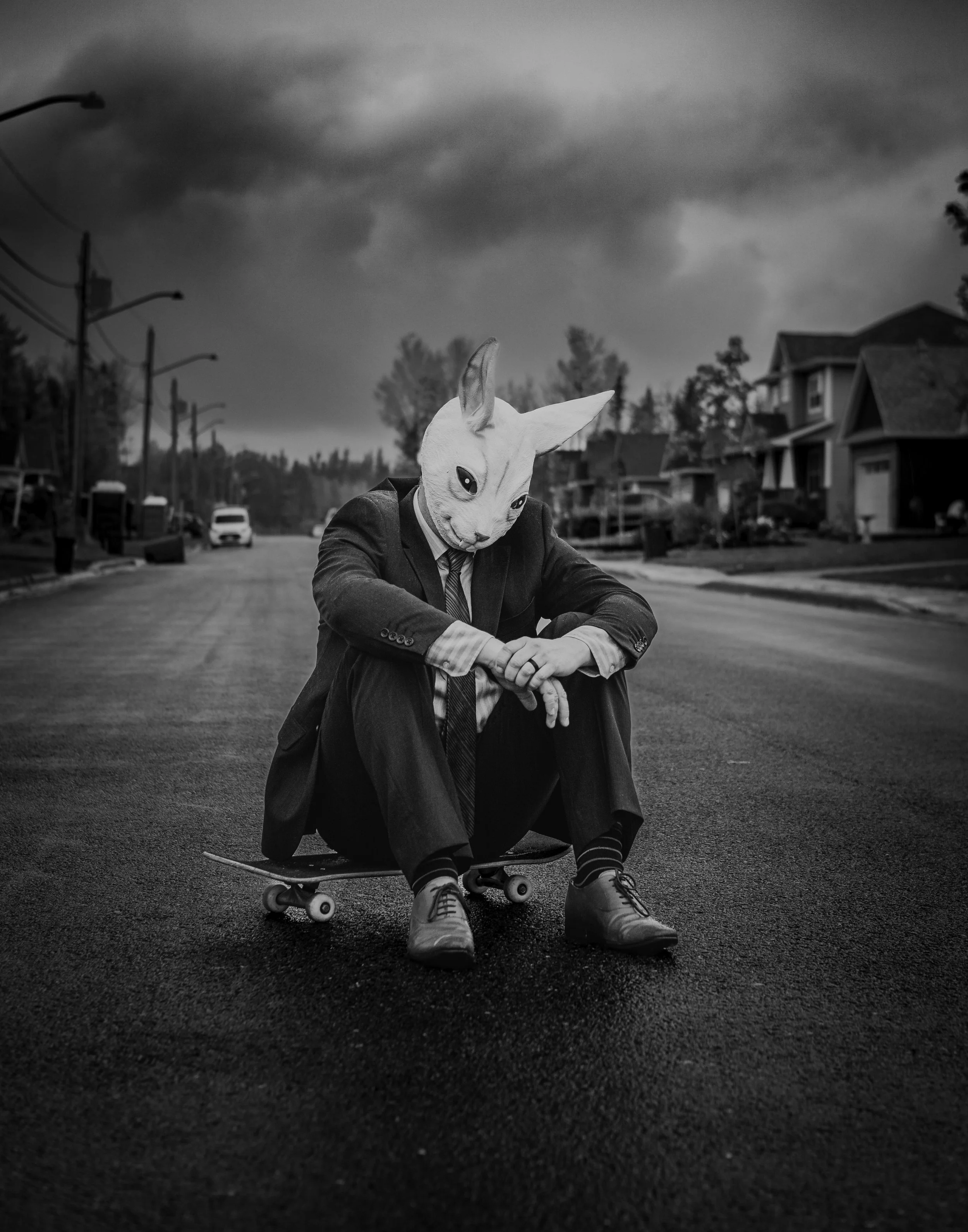 Person in a suit with a rabbit mask sitting on a skateboard in the middle of a street on a cloudy day.