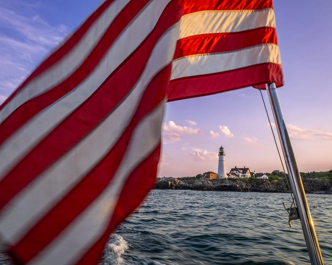 American flag in the foreground on a boat with a lighthouse and house on rocky shore in the background during sunset.