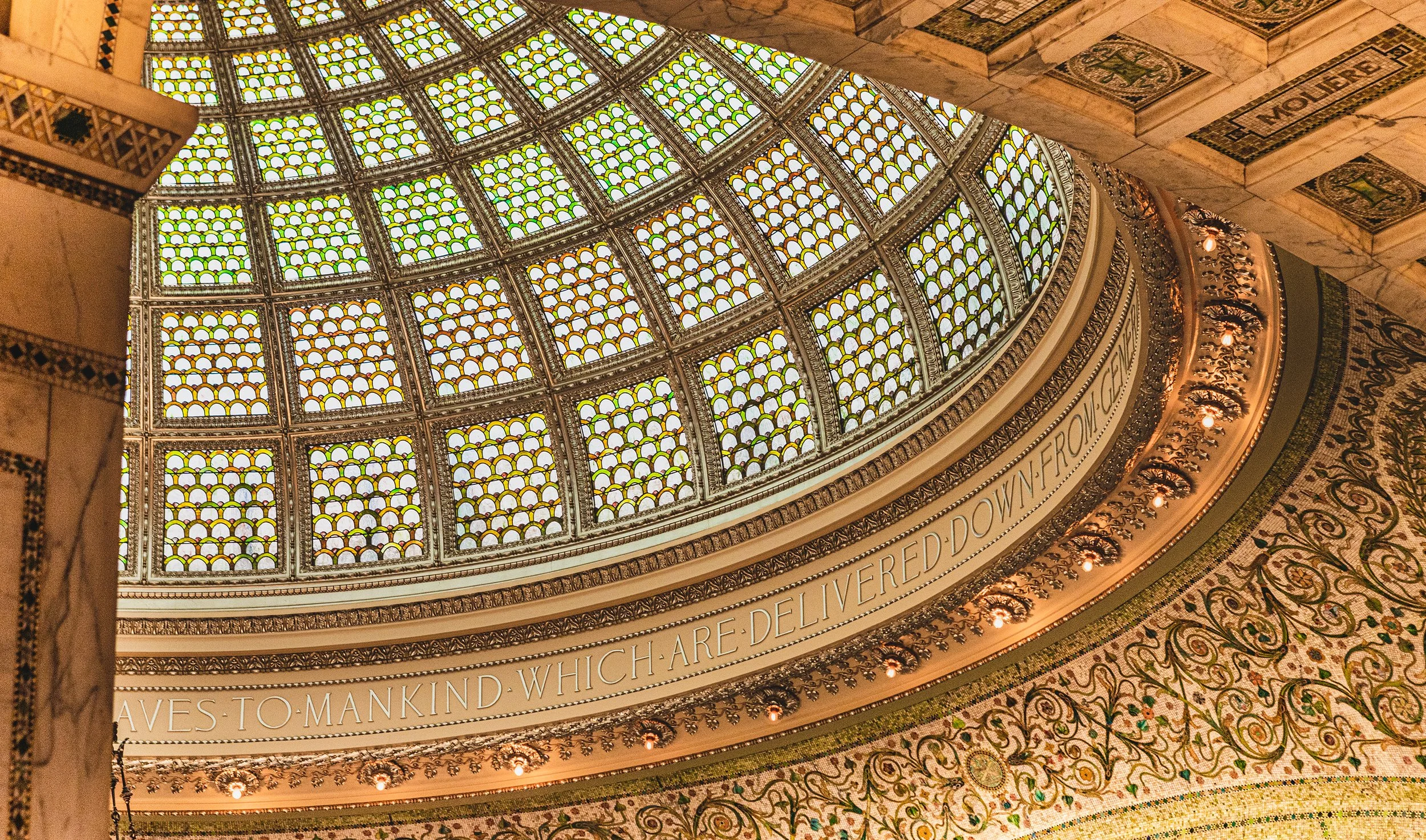 View of a large stained glass dome ceiling with ornate decorative border and a Latin inscription around it.