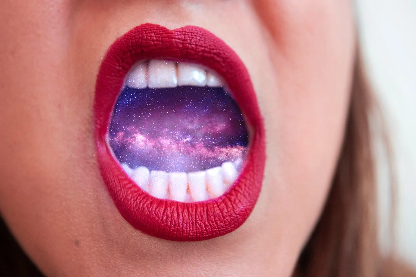 Close-up of a woman's open mouth with red lipstick, showing a galaxy or starry sky inside her mouth where her tongue and the space are visible.