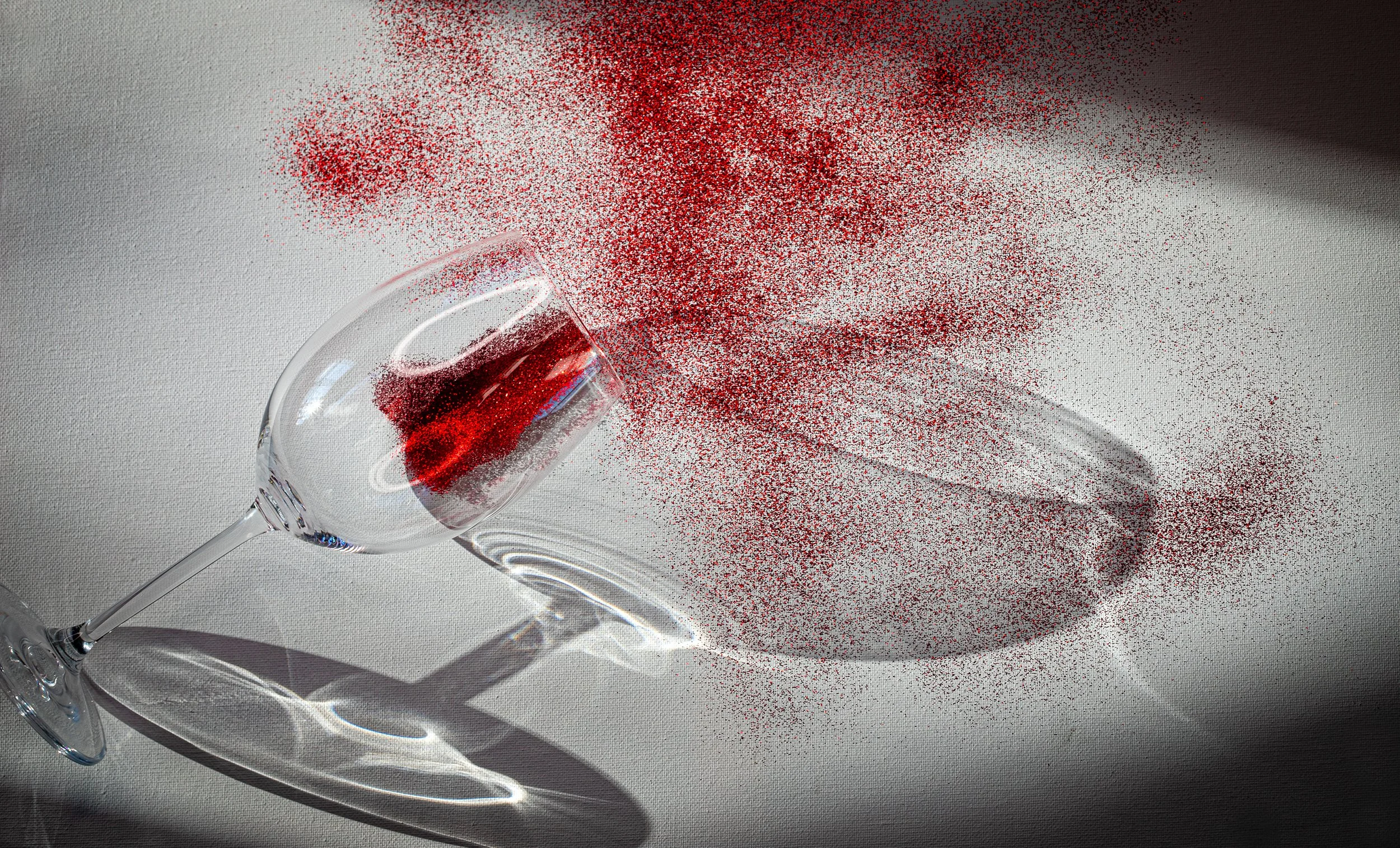 A glass of red wine tipped over on a white surface with red wine spilling and scattering across the surface, creating an abstract pattern of red droplets and a shadow of the glass.
