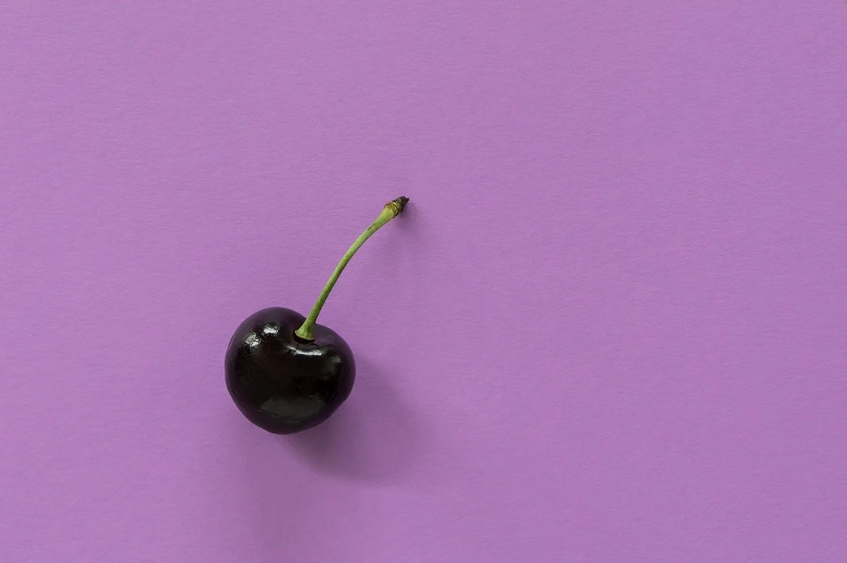 A single black cherry with a green stem on a pink background.
