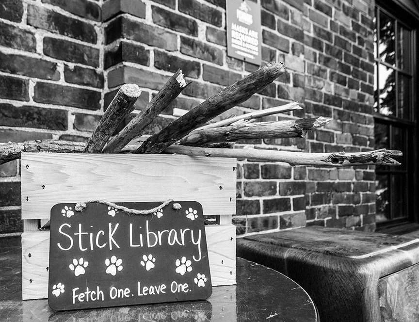 A wooden box filled with large sticks and branches, with a sign that reads 'Stick Library Fetch One. Leave One.' decorated with paw prints, situated on a round table next to a brick wall.