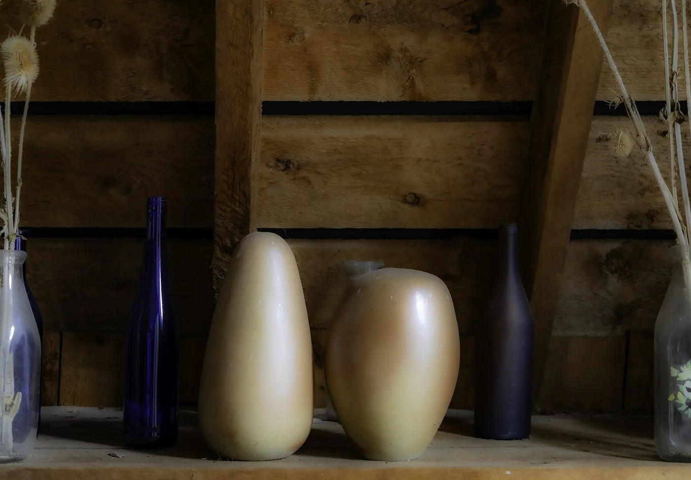 A wooden shelf holding five different vases of varying shapes and colors in a rustic setting.