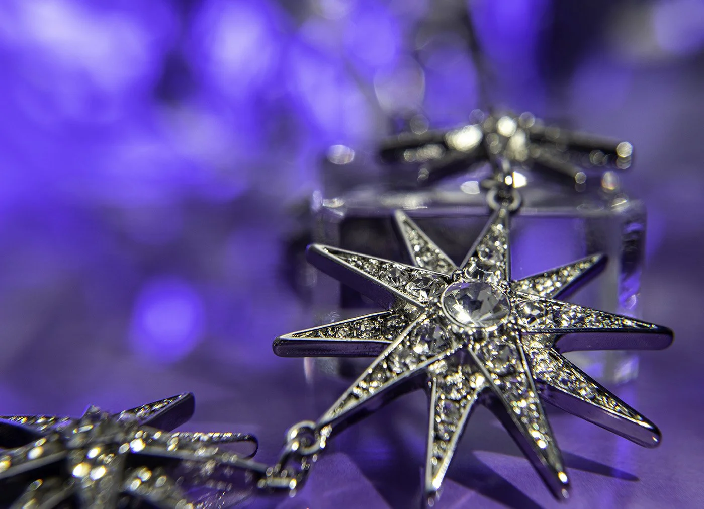 Close-up of a silver star-shaped jewelry pendant with sparkling stones, resting on a purple surface with bokeh lighting in the background.