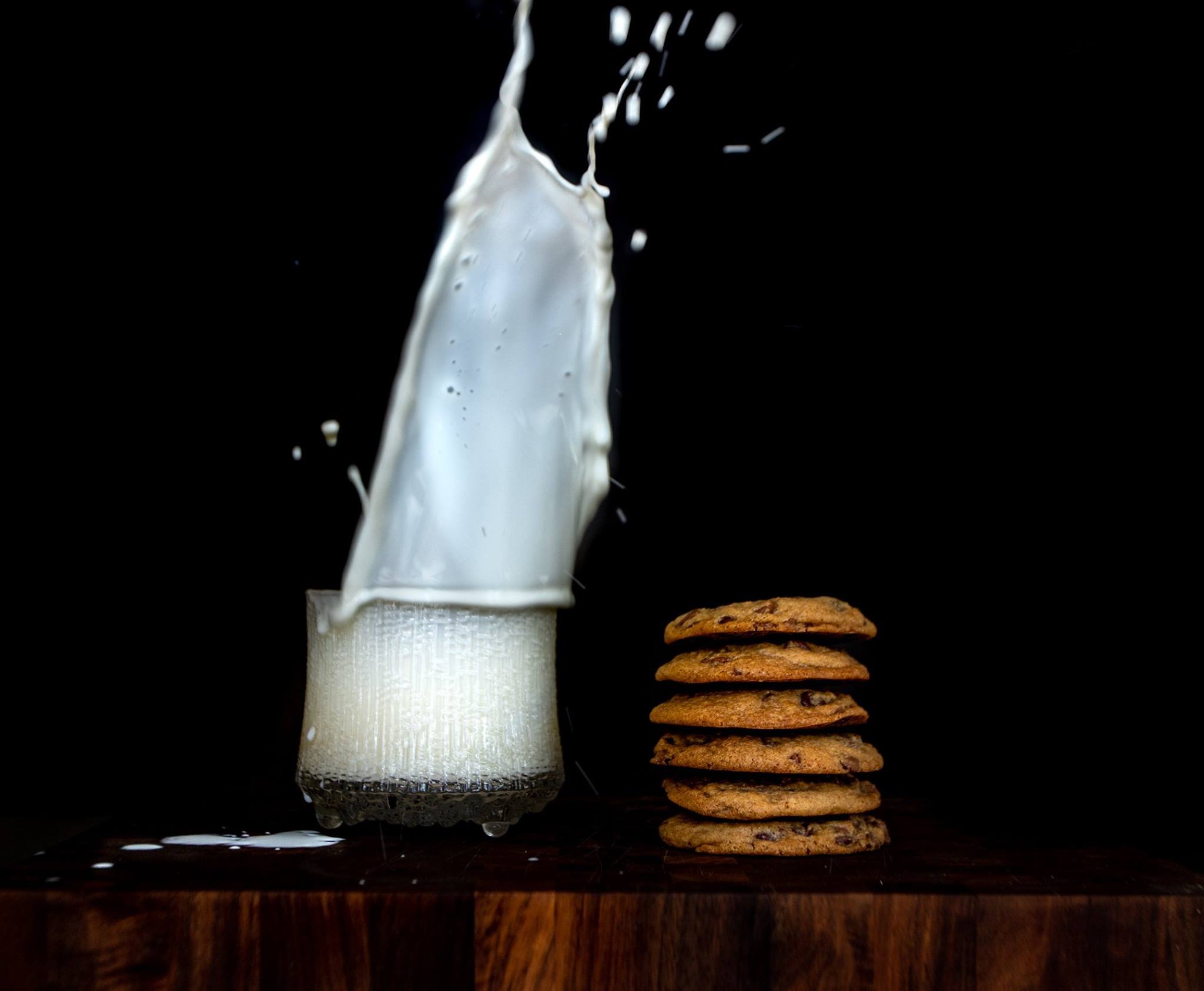 Milky splash falling onto a glass of milk next to a stack of chocolate chip cookies on a wooden surface against a black background.