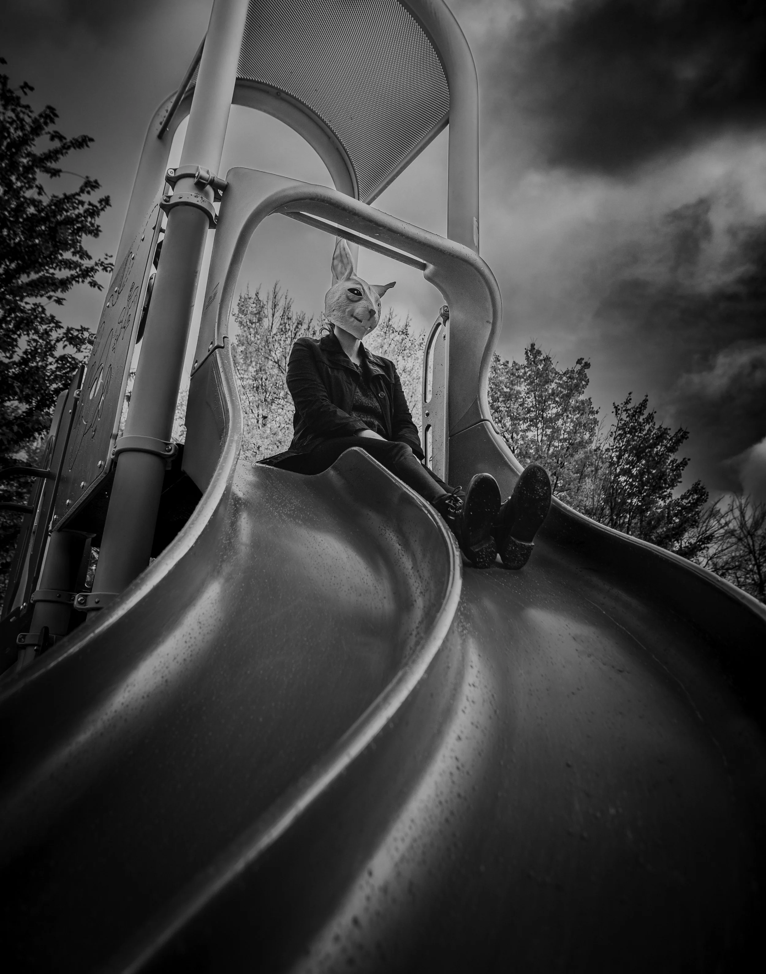 Person sitting at the top of a playground slide with a rabbit mask on their head, taken on a cloudy day.